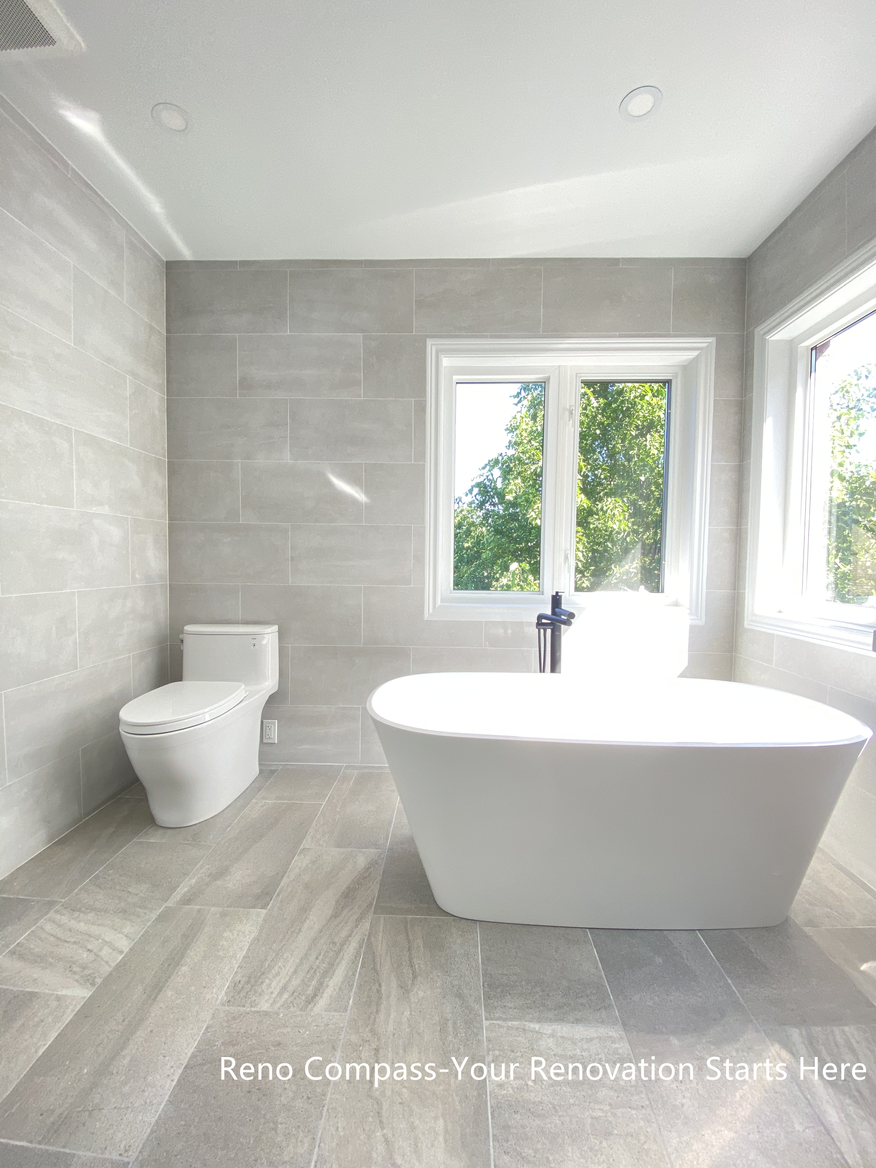 This Aurora bathroom renovation features a freestanding bathtub positioned beside large windows, allowing abundant natural light to enhance the space. Large-format tile flooring and wall finishes create a seamless, spa-inspired environment, while the clean layout maintains a sense of openness and calm. The combination of modern fixtures, soft neutral tones, and daylight results in a bright and relaxing bathing area designed for everyday comfort. Designed and completed by Reno Compass, this bathroom renovation emphasizes clarity, proportion, and long-term livability.