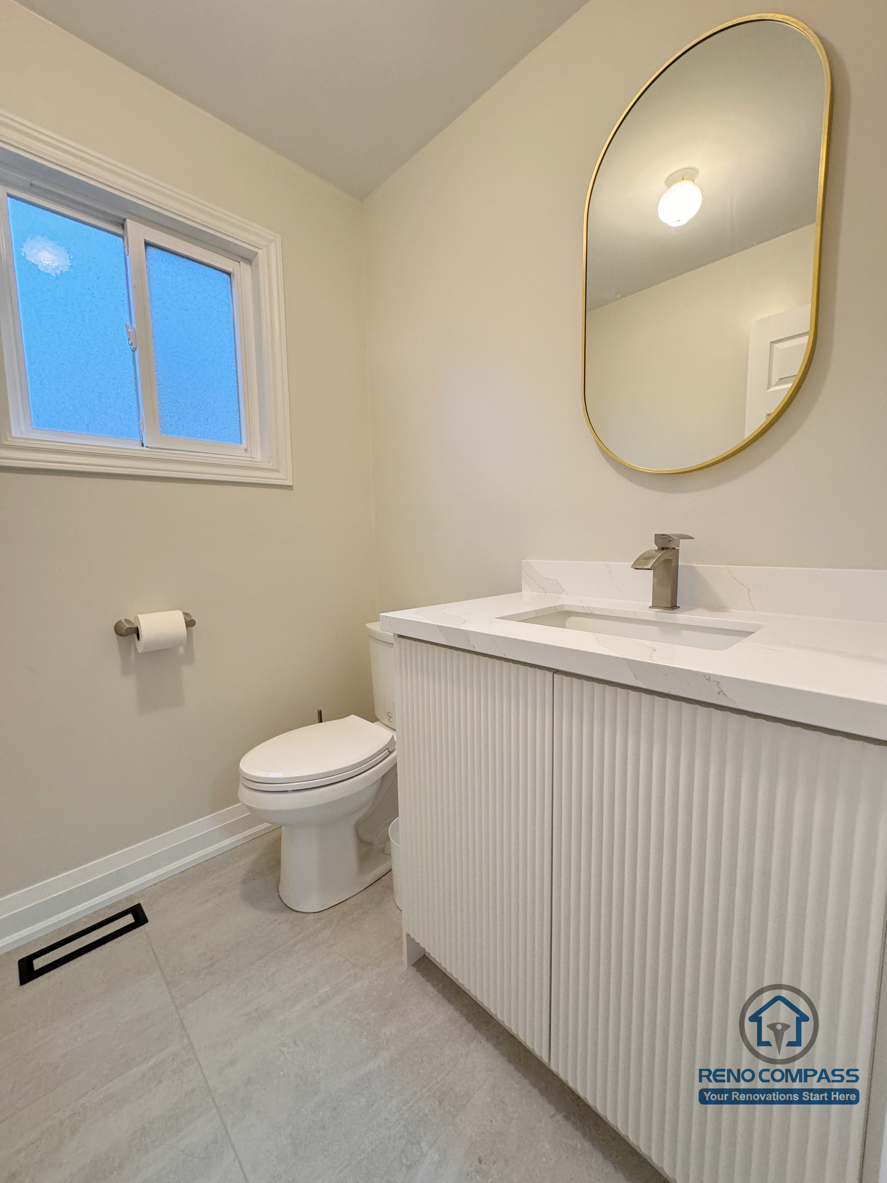 This Mississauga interior renovation features a clean and modern powder room designed with simplicity and functionality in mind. The space includes a custom vanity with vertical panel detailing, a quartz countertop, and a minimalist integrated sink. A gold-framed mirror adds a subtle accent, while neutral tiles and soft lighting create a bright, balanced atmosphere. The layout was carefully planned to maximize comfort and visual openness in a compact space.
