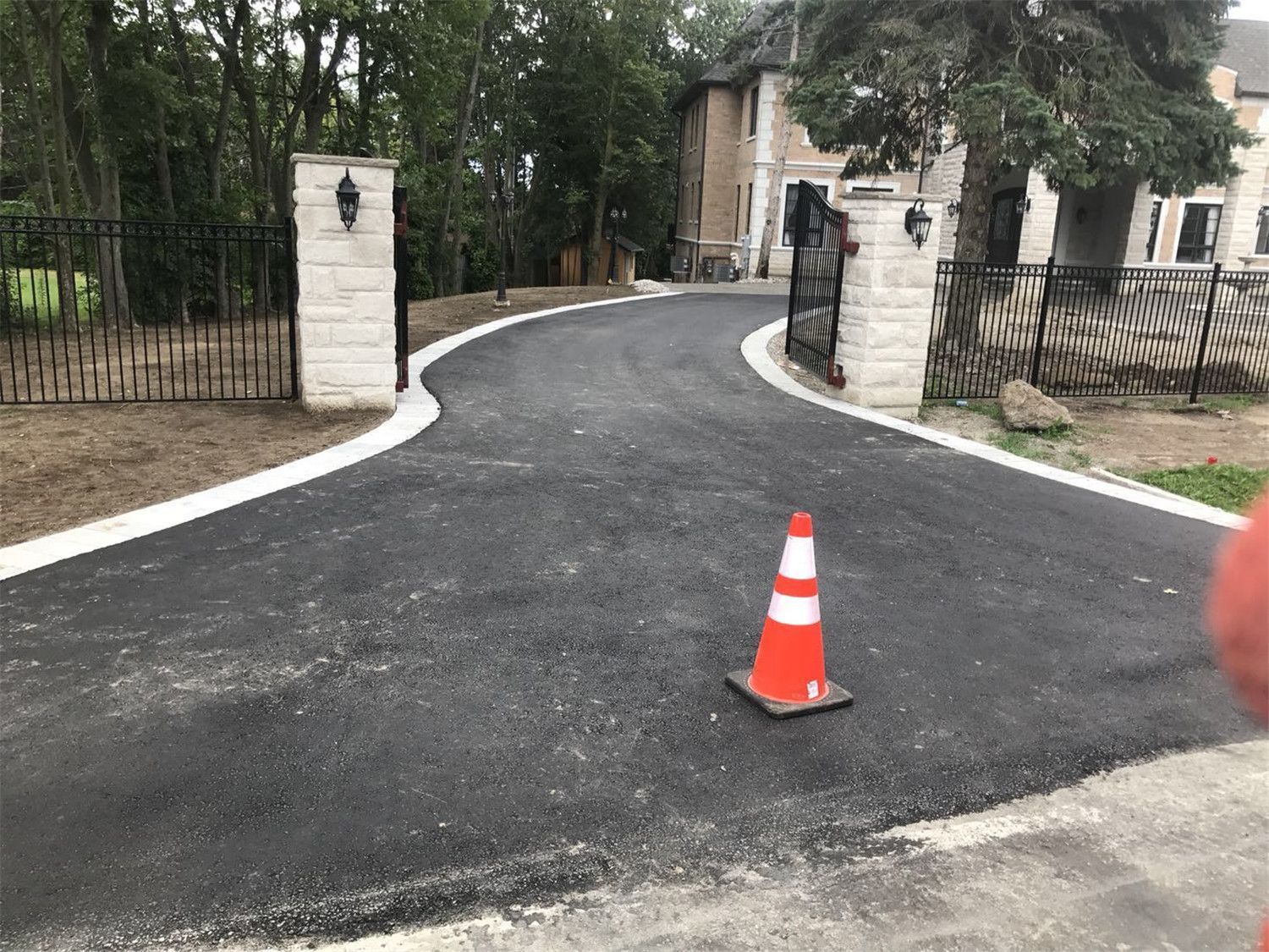 Elegant Toronto driveway landscaping project featuring newly paved asphalt driveway, curved entrance layout, decorative stone gate pillars, and black metal fencing. The design creates a welcoming residential entrance while improving driveway functionality and curb appeal.