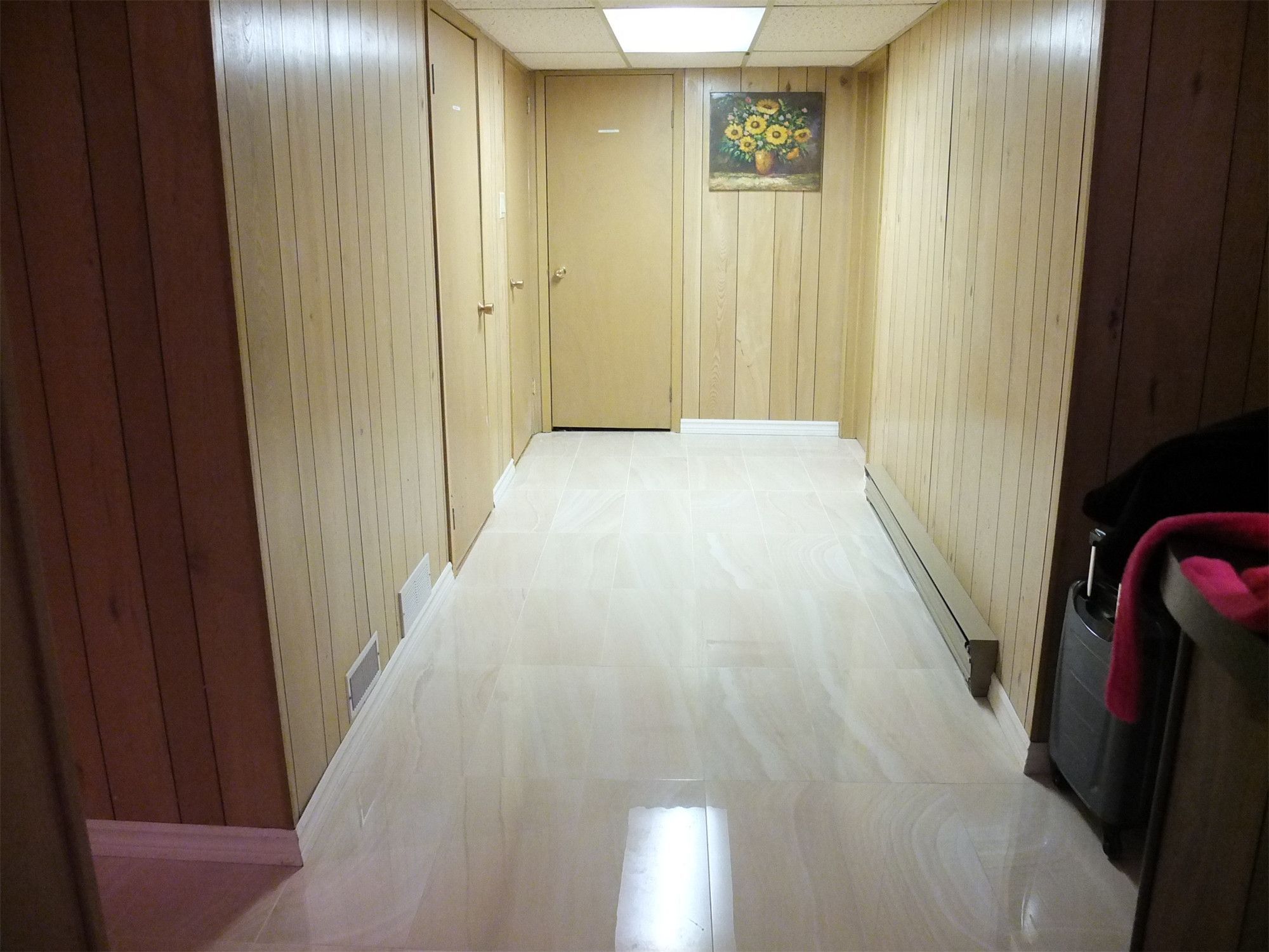 This image presents a finished basement hallway from a North York interior renovation, featuring glossy tile flooring, vertical wood wall panels, and clean transitions along doors and baseboards. The renovation enhances brightness, durability, and spatial continuity, creating a neat and functional circulation area within the basement layout.