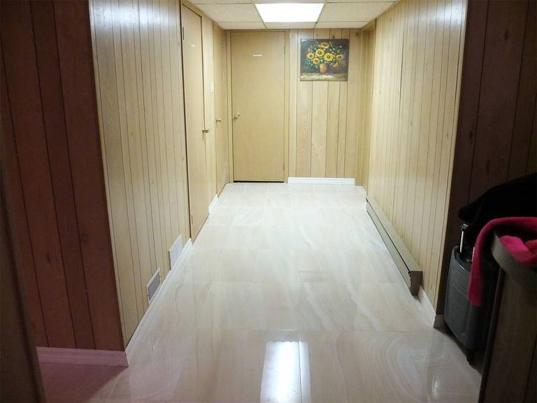 This image presents a finished basement hallway from a North York interior renovation, featuring glossy tile flooring, vertical wood wall panels, and clean transitions along doors and baseboards. The renovation enhances brightness, durability, and spatial continuity, creating a neat and functional circulation area within the basement layout.