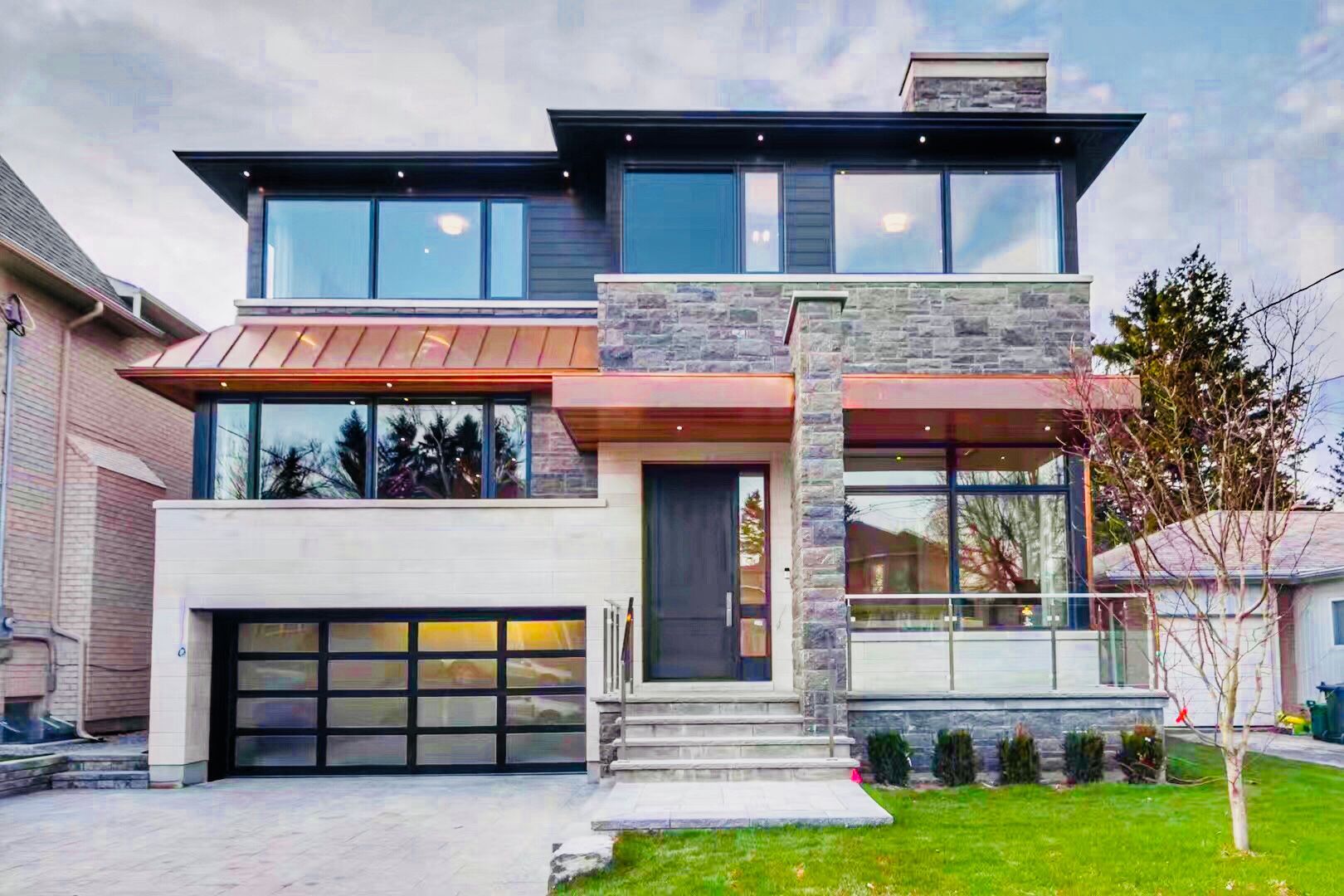 An architectural masterpiece in Toronto featuring a blend of natural fieldstone, limestone, and premium copper roofing accents. This modern custom build showcases expansive floor-to-ceiling windows, a glass-panel garage door, and sophisticated soffit lighting for maximum evening curb appeal.