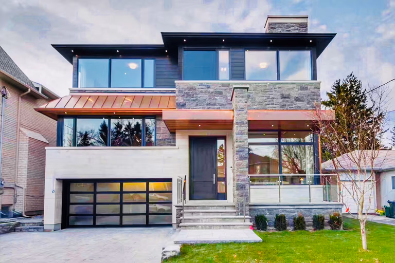 An architectural masterpiece in Toronto featuring a blend of natural fieldstone, limestone, and premium copper roofing accents. This modern custom build showcases expansive floor-to-ceiling windows, a glass-panel garage door, and sophisticated soffit lighting for maximum evening curb appeal.