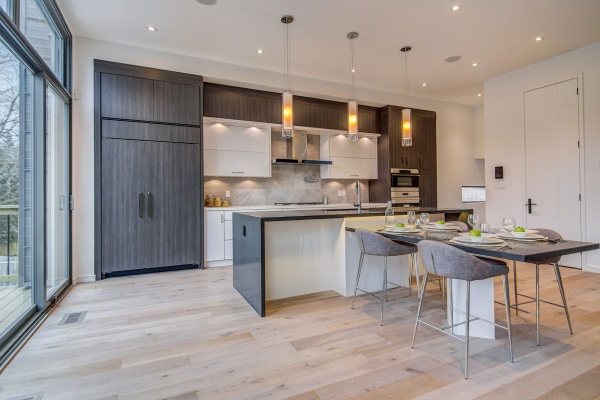 Contemporary Toronto kitchen renovation featuring custom two-tone cabinetry, large central island with seating, quartz countertops, and modern pendant lighting. Floor-to-ceiling windows bring in natural light, enhancing the bright open-concept layout. This modern kitchen was completed by a professional Toronto renovation company, combining functionality, storage, and elegant residential design for upscale living.