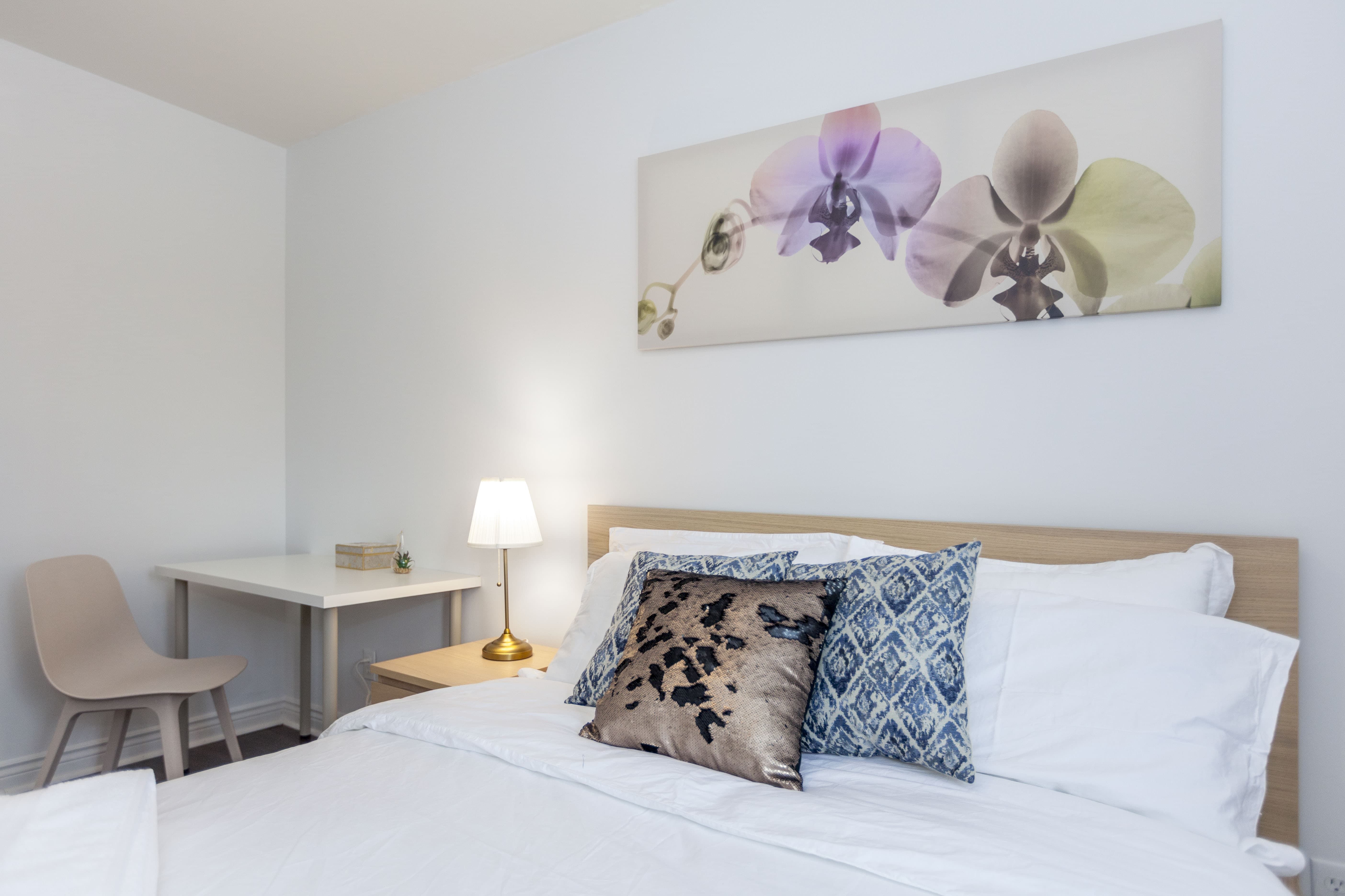 This Toronto interior renovation features a bright and modern bedroom design with a comfortable bed, decorative pillows, bedside lighting, and a compact study desk. The clean white walls and soft furnishings create a calm and inviting atmosphere, making the room both practical and visually appealing. This space reflects the thoughtful planning and finishing quality often seen in a professional Toronto Renovation Company project.