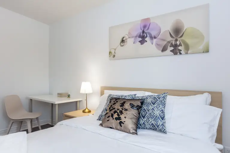 This Toronto interior renovation features a bright and modern bedroom design with a comfortable bed, decorative pillows, bedside lighting, and a compact study desk. The clean white walls and soft furnishings create a calm and inviting atmosphere, making the room both practical and visually appealing. This space reflects the thoughtful planning and finishing quality often seen in a professional Toronto Renovation Company project.