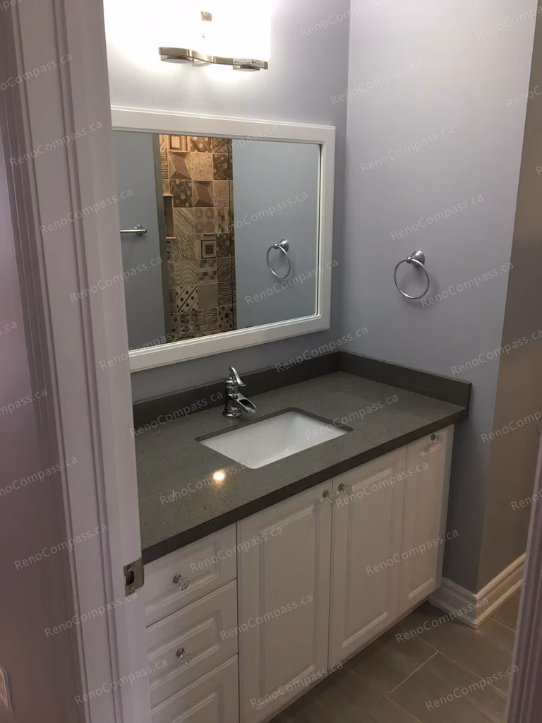 This Markham interior renovation highlights a modern bathroom vanity upgrade featuring custom white cabinetry, a durable stone countertop, and a sleek integrated sink. The large framed mirror and updated lighting enhance brightness and functionality, creating a clean and elegant bathroom space. Completed by Reno Compass, a trusted Toronto renovation company specializing in bathroom and interior home renovations.