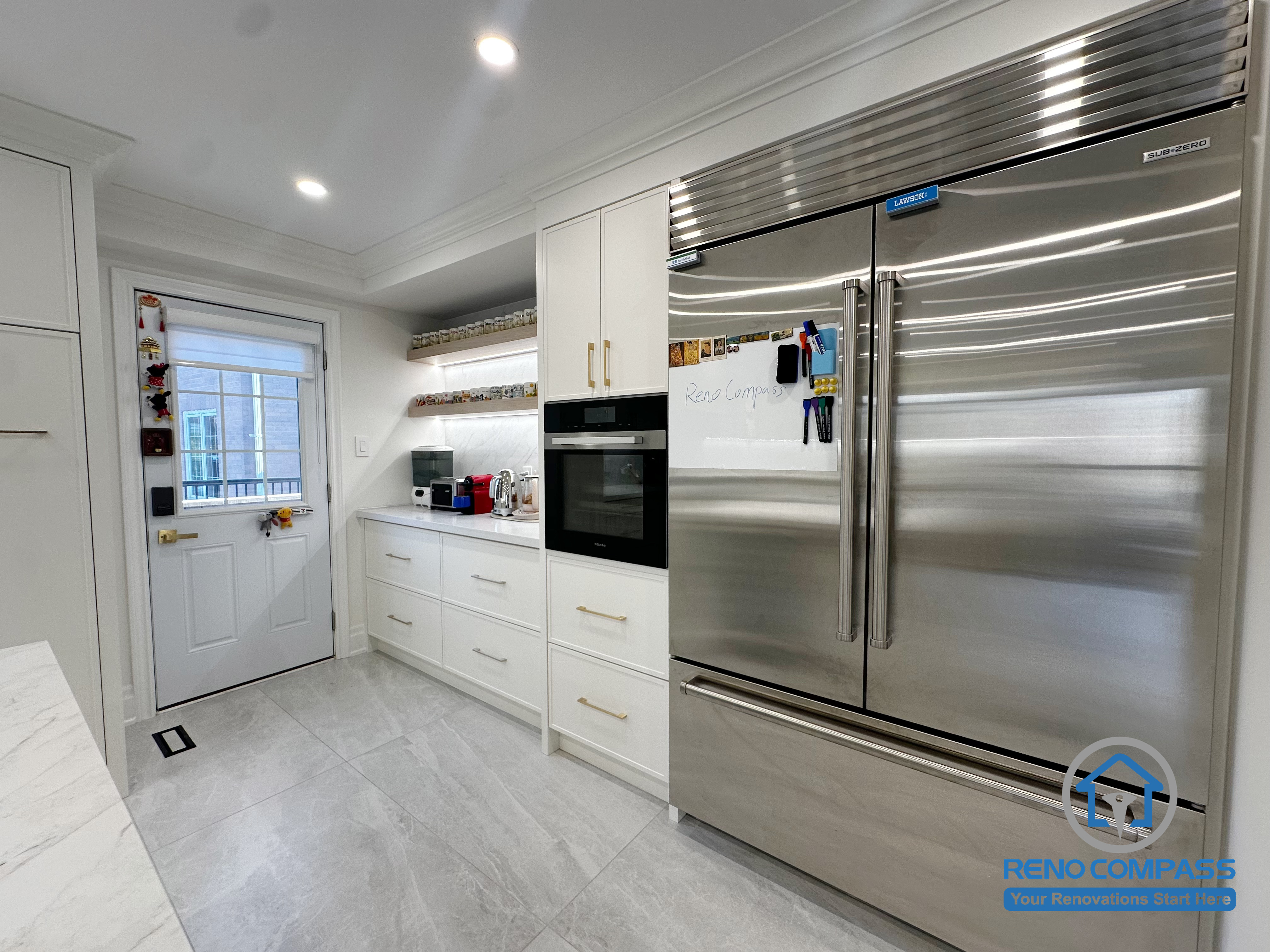 This kitchen renovation highlights custom white cabinetry designed to integrate built-in appliances and maximize storage efficiency. A stainless steel refrigerator, wall oven, and open shelving are thoughtfully arranged to support everyday cooking needs while maintaining a clean, streamlined appearance. The layout balances functionality with a modern, polished finish.