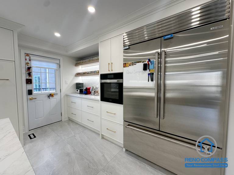 This kitchen renovation highlights custom white cabinetry designed to integrate built-in appliances and maximize storage efficiency. A stainless steel refrigerator, wall oven, and open shelving are thoughtfully arranged to support everyday cooking needs while maintaining a clean, streamlined appearance. The layout balances functionality with a modern, polished finish.