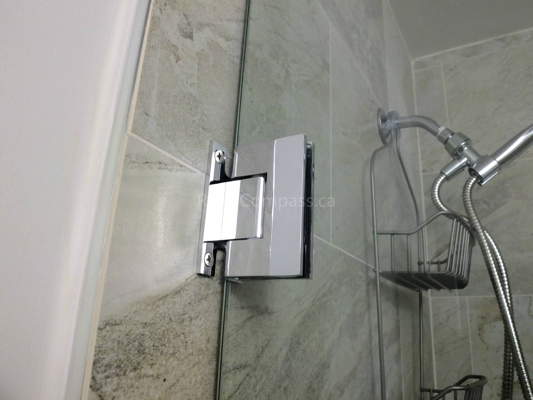 This image captures a close-up of a frameless glass shower door hinge installed during a North York interior renovation project. The solid chrome hinge is precisely mounted into tiled walls, ensuring smooth door operation, long-term durability, and a clean, minimalist appearance. Combined with marble-look wall tiles and modern shower fixtures, the detail reflects careful hardware selection and professional installation by a trusted Toronto renovation company.