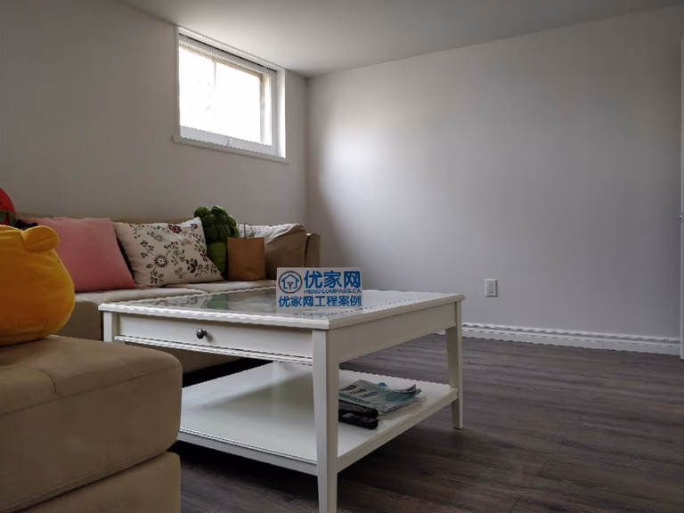This Scarborough basement renovation showcases a comfortable finished living area featuring modern vinyl flooring, freshly painted walls, and upgraded baseboards. The space is styled with a sofa and coffee table to create a cozy lounge area, while the basement window allows natural light to brighten the room. This functional design transforms the basement into a welcoming relaxation space perfect for family gatherings or everyday living.