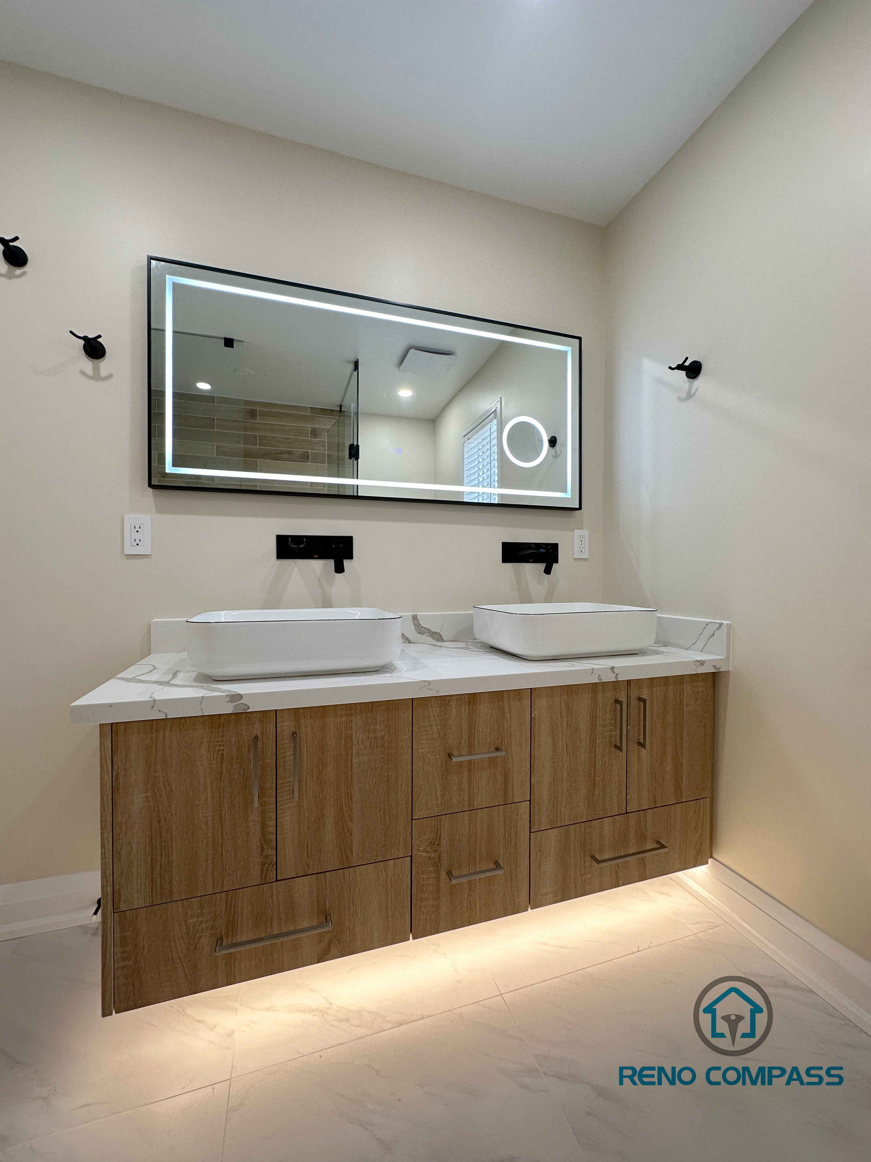 Modern bathroom renovation in Vaughan featuring a custom double vanity with wood-finish cabinetry, quartz countertop, and two vessel sinks. The vanity is paired with a large backlit LED mirror and matte black wall-mounted faucets, creating a clean and contemporary look. Integrated under-cabinet lighting enhances both functionality and visual warmth. This bathroom design highlights precise installation and material coordination by a professional Toronto renovation company focused on modern comfort and refined details.