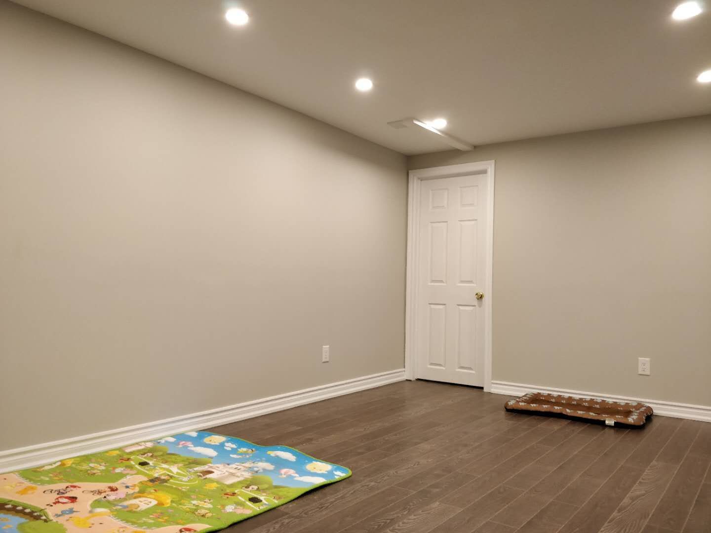 This image showcases a completed basement renovation project in Scarborough featuring a clean, modern layout. The space includes dark hardwood flooring, neutral wall finishes, and recessed pot lighting that enhances brightness and visual openness. The minimalist design creates a versatile area suitable for a family lounge, children's playroom, or multi-purpose living space. Completed by Reno Compass Inc., this basement renovation emphasizes quality craftsmanship, clean detailing, and a comfortable interior environment.