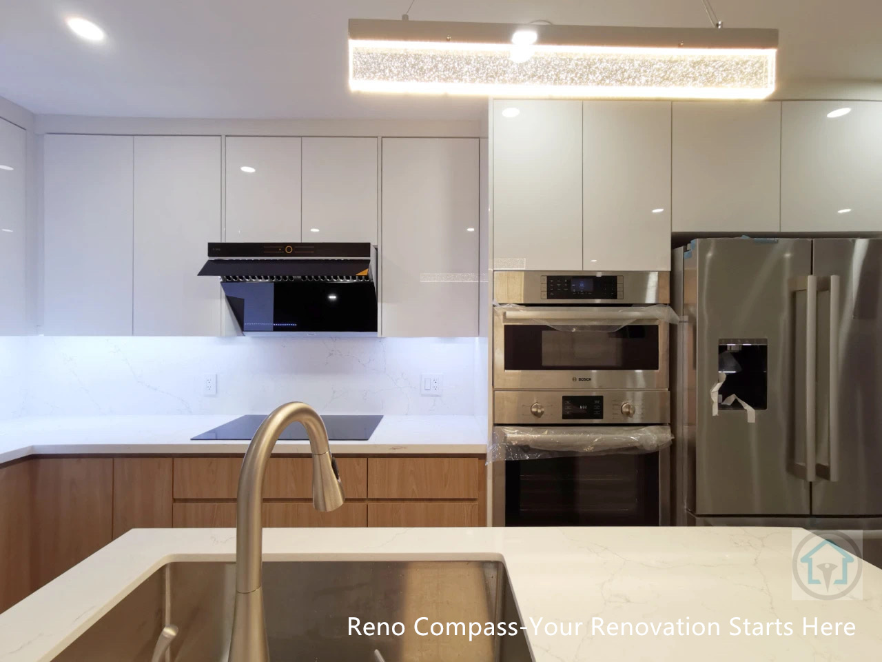 This image highlights the completed open-concept kitchen from a Richmond Hill interior renovation project. The design centers around a custom-built kitchen island with an integrated sink and clean waterfall-style countertop, creating a strong visual focal point while enhancing everyday functionality. Full-height glossy cabinetry maximizes storage and maintains a seamless, modern appearance, while the combination of warm wood base cabinets and white upper cabinets adds balance and depth to the space.

Built-in stainless steel appliances, including a wall oven, microwave, and refrigerator, are neatly integrated to keep the layout organized and clutter-free. The statement linear pendant light above the island provides both task lighting and a refined architectural accent, complementing the recessed ceiling lights for layered illumination. A sleek range hood and continuous backsplash further reinforce the minimalist, contemporary aesthetic.

Designed and constructed by Reno Compass Inc., this kitchen renovation reflects the quality and detail expected from a professional Toronto renovation company, delivering a bright, functional, and timeless kitchen space tailored for modern living.