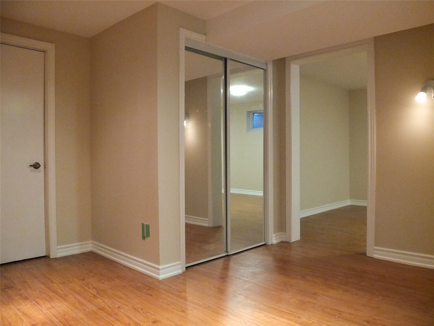 This Markham basement renovation features a finished bedroom area with warm neutral wall paint, durable laminate flooring, and sliding mirror closet doors. The mirrored closet helps enhance brightness and create a sense of larger space, while the clean wall finishing, trim work, and lighting provide a comfortable and functional living area within the basement renovation.