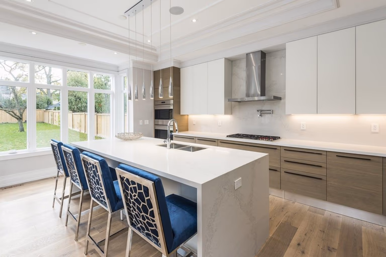 This Newmarket kitchen renovation showcases a bright open-concept layout completed by a professional Toronto renovation company. The space features a large waterfall-edge quartz island with seating, custom two-tone cabinetry, built-in stainless steel appliances, and modern pendant lighting. Expansive windows bring in natural light and connect the kitchen to the backyard, creating a warm and functional family space. The renovation enhances workflow, storage, and modern aesthetics, delivering a high-end kitchen design ideal for everyday living and entertaining in Newmarket and across the GTA.