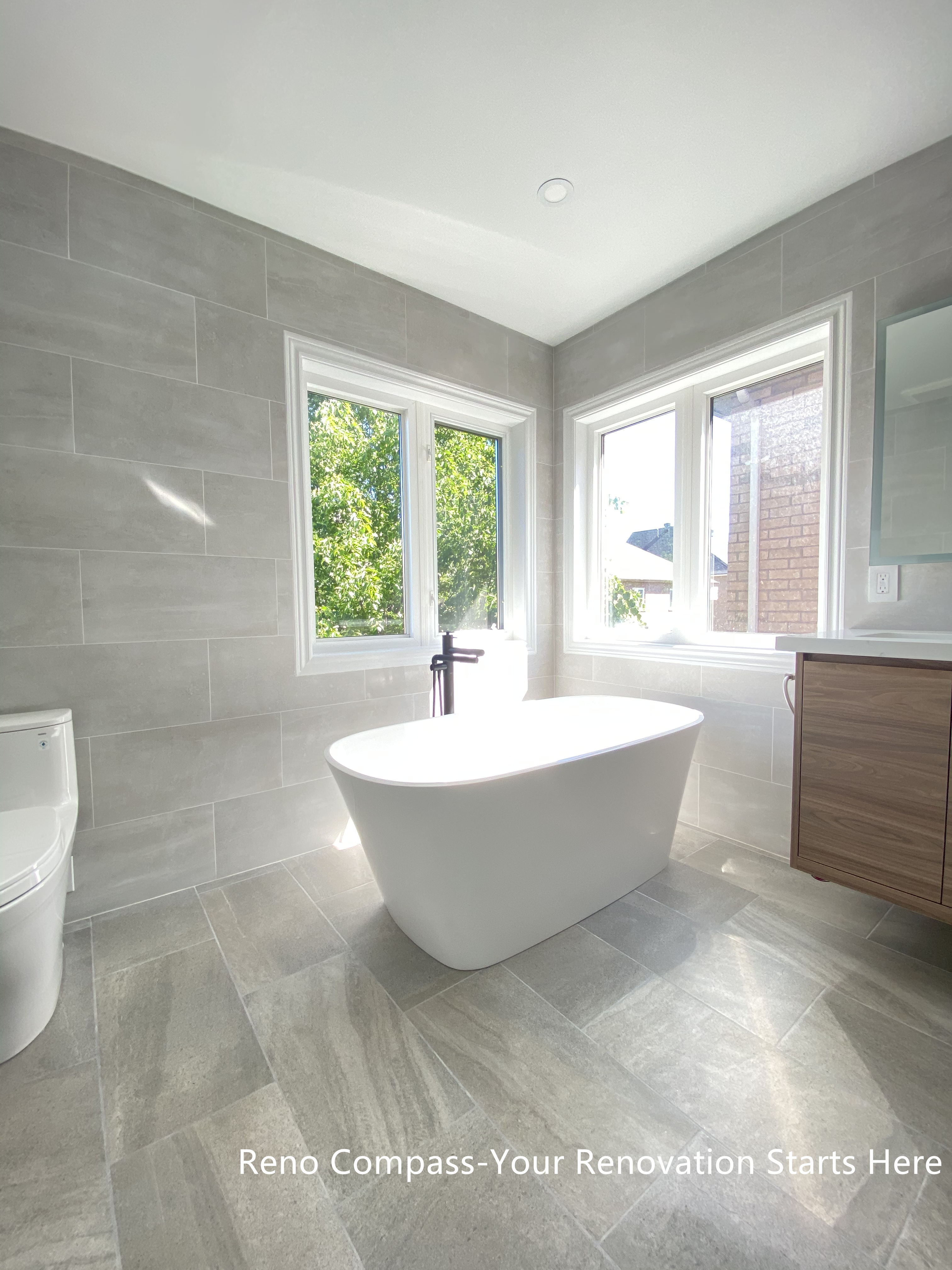 This Aurora bathroom renovation highlights a freestanding bathtub positioned beside corner windows, allowing natural light to fill the space throughout the day. Large-format tile flooring and wall finishes create a seamless, modern backdrop, while the clean tub silhouette and minimalist fixtures reinforce a calm, spa-inspired atmosphere. The layout maintains generous circulation and visual openness, making the bathing area both functional and relaxing. Designed and completed by Reno Compass, this bathroom renovation emphasizes comfort, proportion, and long-term usability.