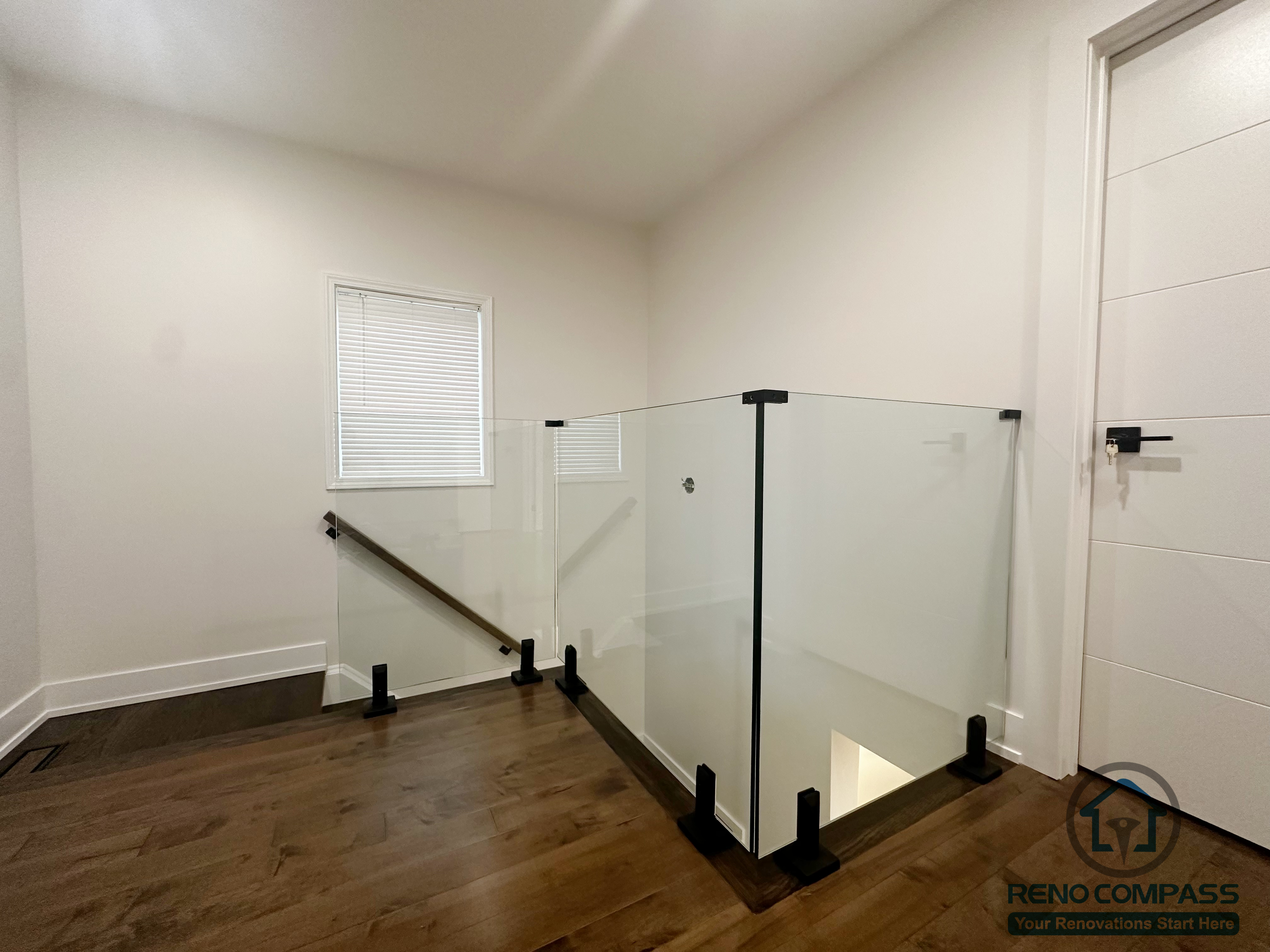 This Oakville interior renovation features a clean upper-level stair landing enclosed with a glass guardrail for safety and visual openness. The transparent railing allows natural light to travel between floors while maintaining a clear boundary along the stair opening. Warm wood flooring and simple wall finishes create a calm transition space that connects private rooms and circulation areas with a modern, understated aesthetic.