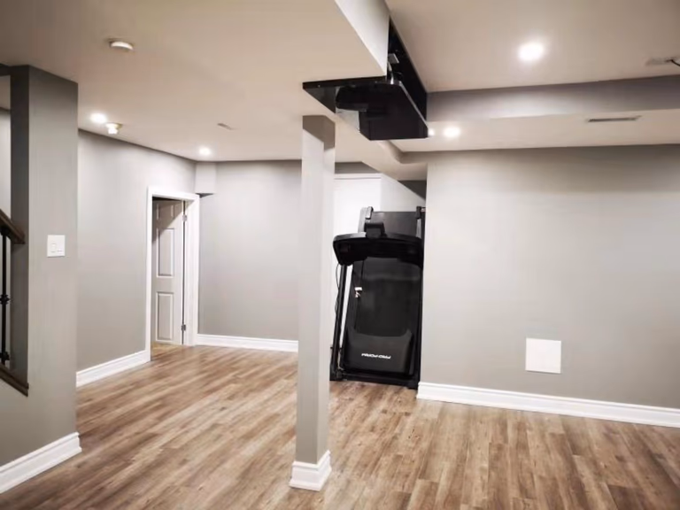 This finished basement renovation in Mississauga features durable vinyl flooring, recessed pot lights, and a clean open-concept layout with structural columns. The space is designed for multi-functional use, including a home gym area, showcasing the expertise of a professional Toronto renovation company in delivering modern, practical basement transformations.