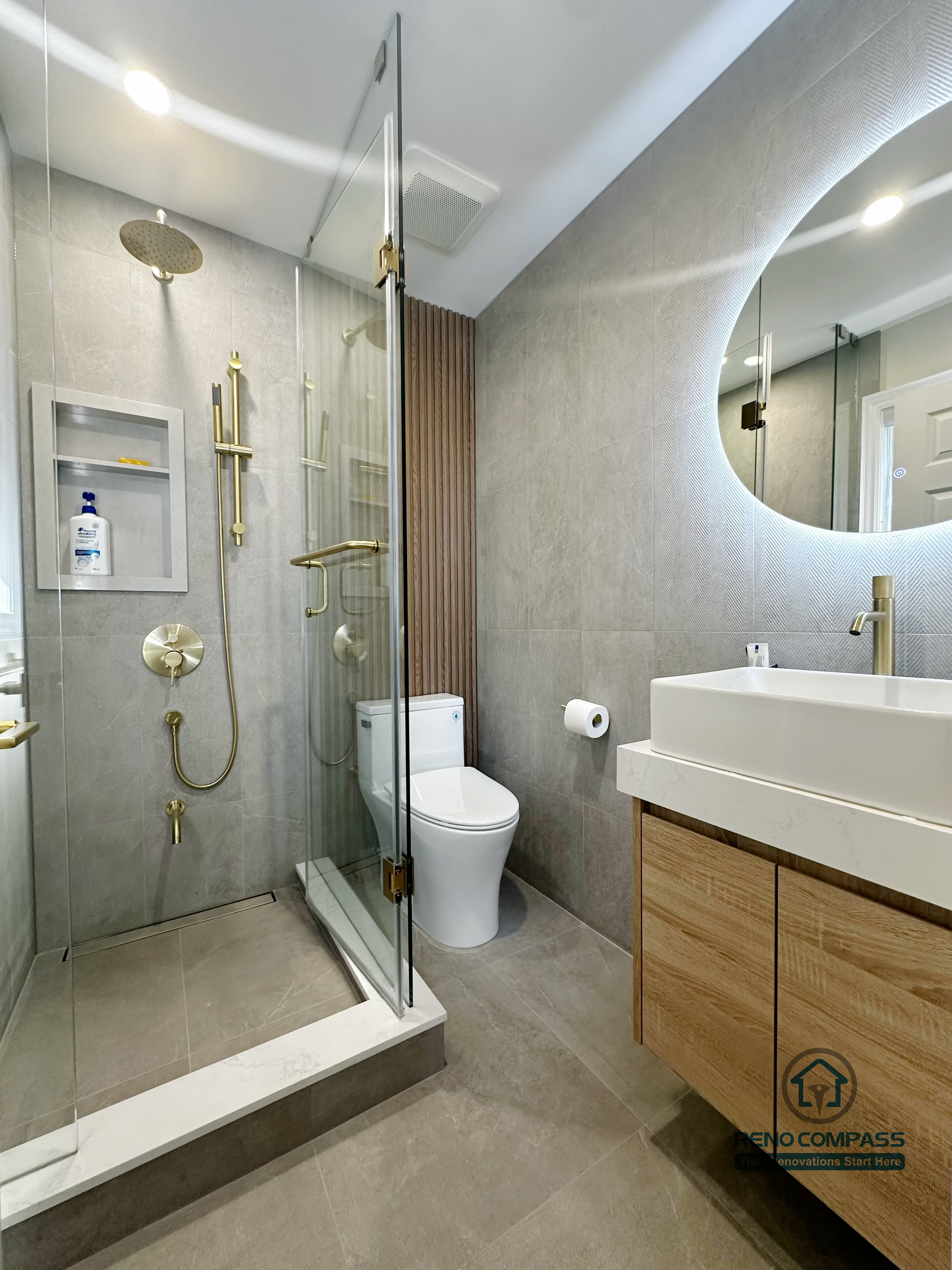 This image highlights a detailed bathroom renovation completed in North York, featuring a walk-in glass shower with built-in wall niche and brushed gold shower fixtures. The space includes a floating wood vanity with integrated storage, a modern toilet, and large-format tile flooring that enhances durability and visual consistency. Frameless glass panels and warm metallic accents create a clean, contemporary bathroom design focused on comfort, functionality, and refined detailing for North York homes.