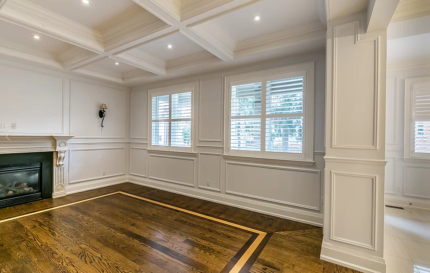 This Richmond Hill interior renovation highlights a refined living space with a coffered ceiling, custom wainscoting, and detailed hardwood flooring with inlay borders. Large windows with plantation shutters enhance natural light, while classic millwork and clean architectural lines create a timeless, elegant interior renovation result.