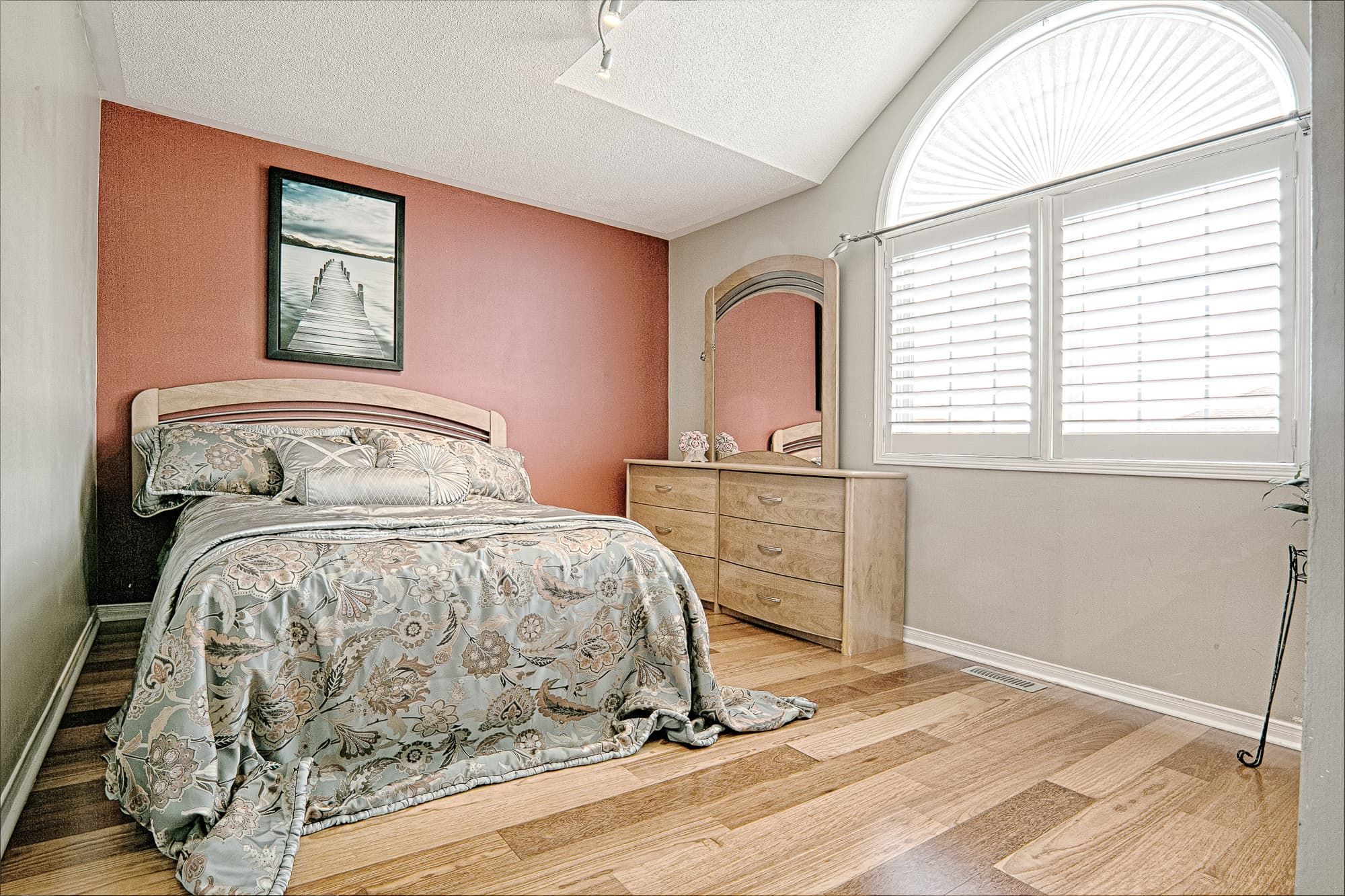 This cozy bedroom interior features a warm accent wall, natural hardwood flooring, and soft patterned bedding that create an inviting and comfortable atmosphere. The coordinated dresser set, arched mirror, and large window with plantation shutters enhance both functionality and natural light. A beautiful example of warm, minimalist bedroom interior design ideal for homeowners seeking stylish and comfortable home renovation inspiration in Toronto and the GTA.