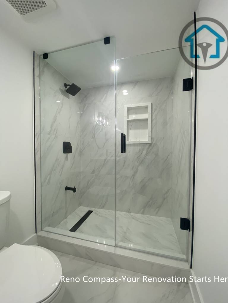 This image showcases a modern basement bathroom renovation in Newmarket completed by a professional Toronto renovation company. The walk-in shower features full-height marble-look tiles, a frameless glass enclosure with black hardware, built-in niche shelving, and a linear floor drain. Clean white finishes and contemporary fixtures create a bright, low-maintenance, and functional basement bathroom ideal for rental units or family use.