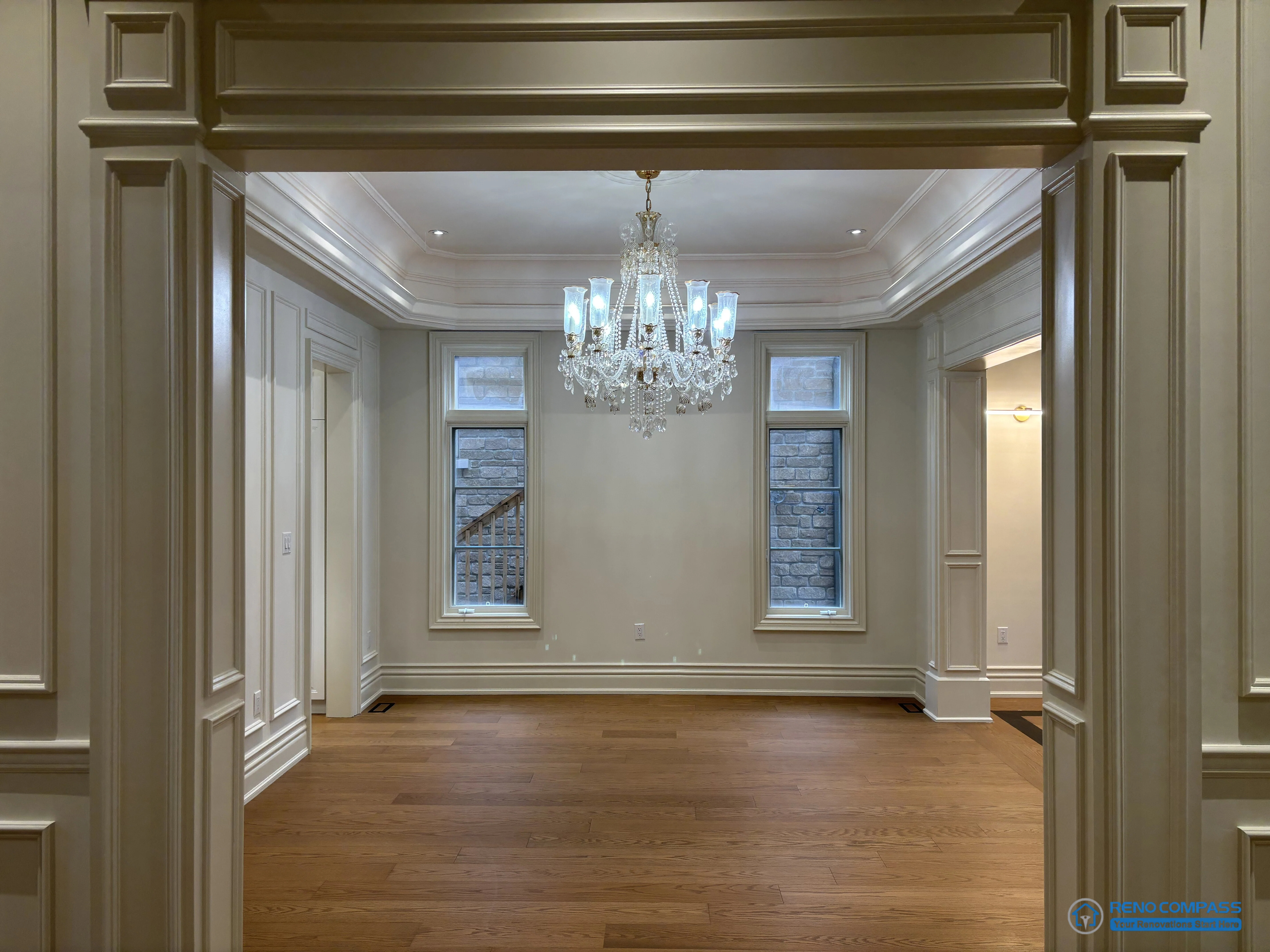 This image captures a European-style living room framed by a classically detailed doorway, emphasizing axial alignment and architectural proportion. The doorway functions as a structural frame, guiding the view toward the central crystal chandelier and symmetrically arranged windows beyond. Crown moulding, wall panelling, and ceiling detailing establish a clear architectural hierarchy, while hardwood flooring grounds the space and reinforces continuity. Rather than relying on decoration, the composition highlights disciplined planning, balanced symmetry, and refined craftsmanship within a high-end Richmond Hill interior renovation rooted in traditional European design principles.