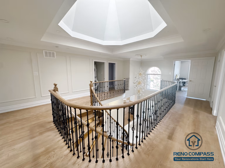 This image showcases a combined flooring and stair renovation completed in North York, featuring hardwood flooring on the upper level and a curved stair railing overlooking the main floor. The staircase is framed with decorative iron balusters and a smooth hardwood handrail, creating continuity between levels. Natural light from the overhead skylight enhances the warmth of the flooring and highlights the architectural detailing. This renovation demonstrates seamless coordination between flooring installation and stair design, improving both visual flow and spatial connection within the home.