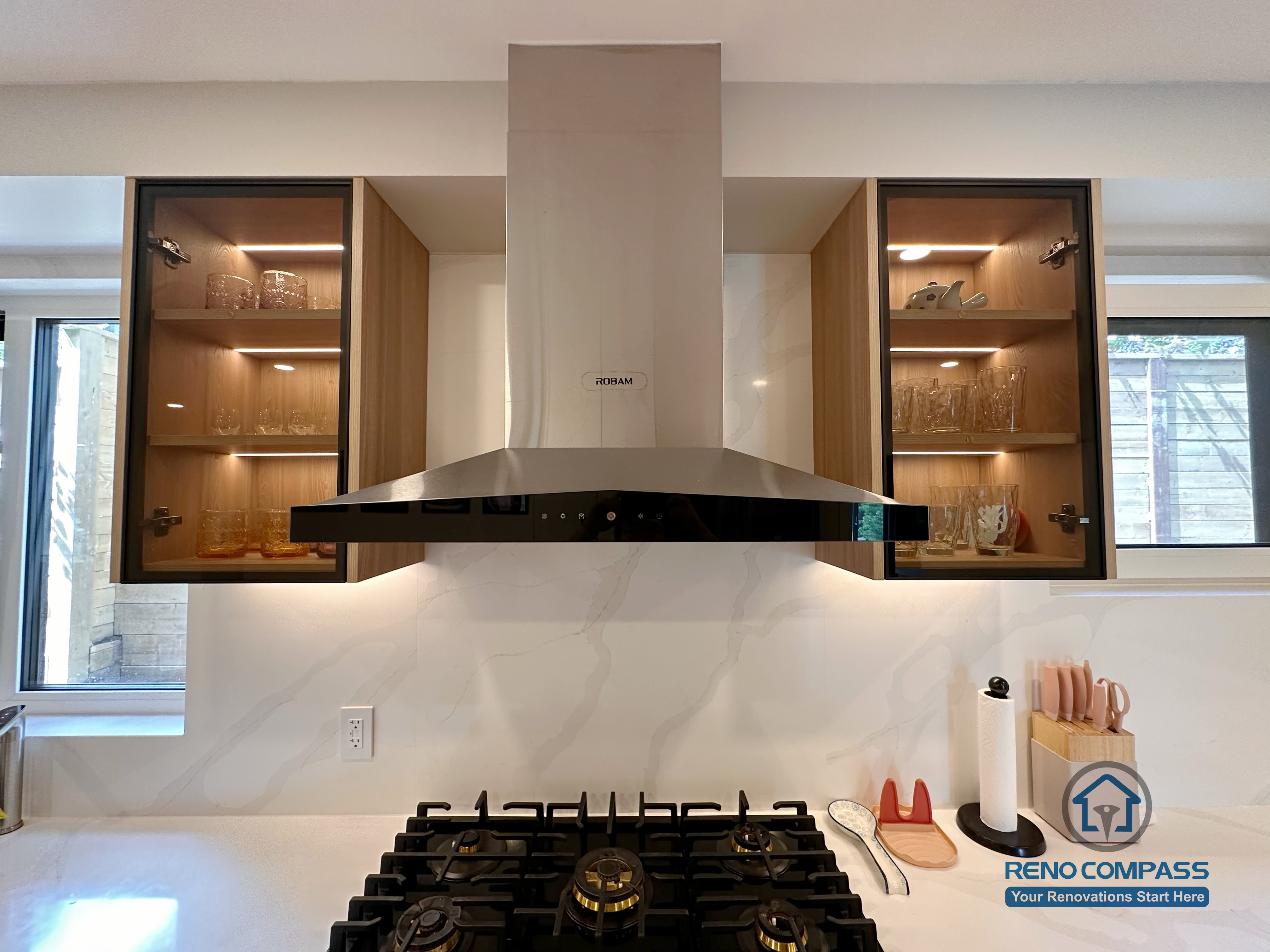 A modern custom kitchen renovation in Toronto showcasing glass display cabinets with integrated LED lighting and a sleek stainless steel range hood. This bespoke cabinetry design combines functionality and visual balance, highlighting fine craftsmanship and thoughtful layout by a professional Toronto renovation company.