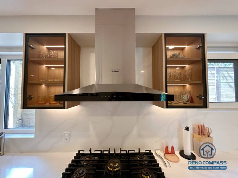 A modern custom kitchen renovation in Toronto showcasing glass display cabinets with integrated LED lighting and a sleek stainless steel range hood. This bespoke cabinetry design combines functionality and visual balance, highlighting fine craftsmanship and thoughtful layout by a professional Toronto renovation company.