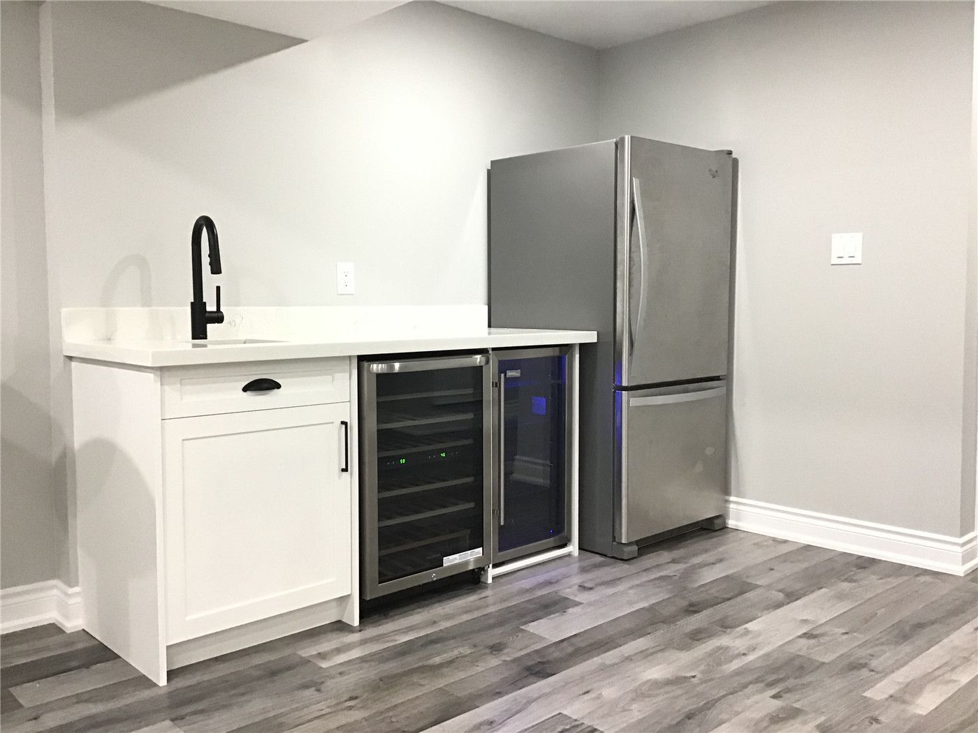 Elegant basement wet bar installation in this Newmarket renovation featuring white shaker cabinetry, quartz countertop, matte black faucet, built-in beverage fridge, and stainless steel refrigerator. A perfect addition for entertaining guests and enhancing basement functionality.