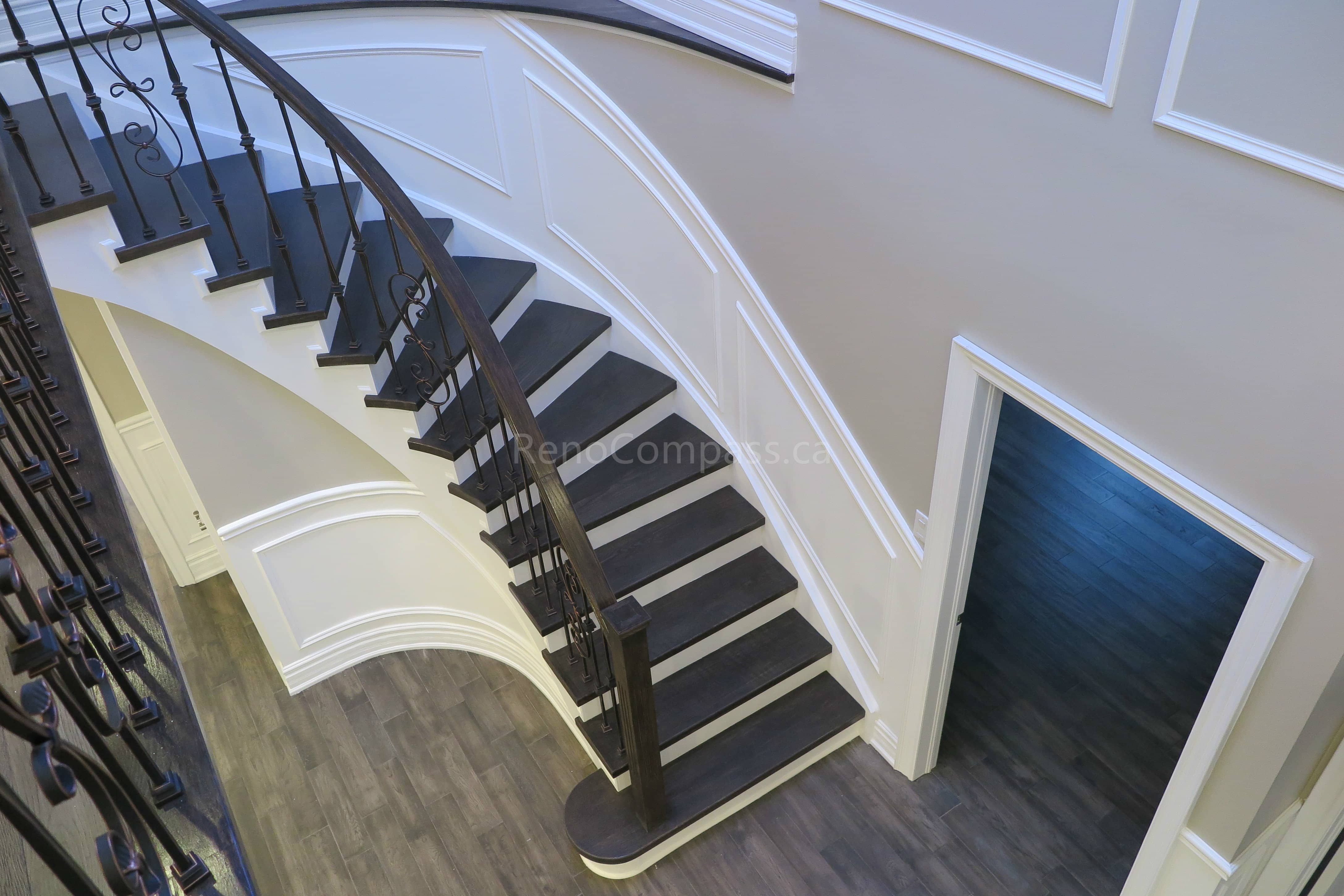 This Markham interior renovation features a stunning custom curved staircase with dark wood treads, white risers, and elegant wrought iron railings. Detailed wall panel molding and modern flooring enhance the sophisticated architectural design, creating a luxurious focal point within the home. Completed by Reno Compass, a professional renovation contractor specializing in custom staircases and high-end interior renovations across Markham and the Greater Toronto Area.