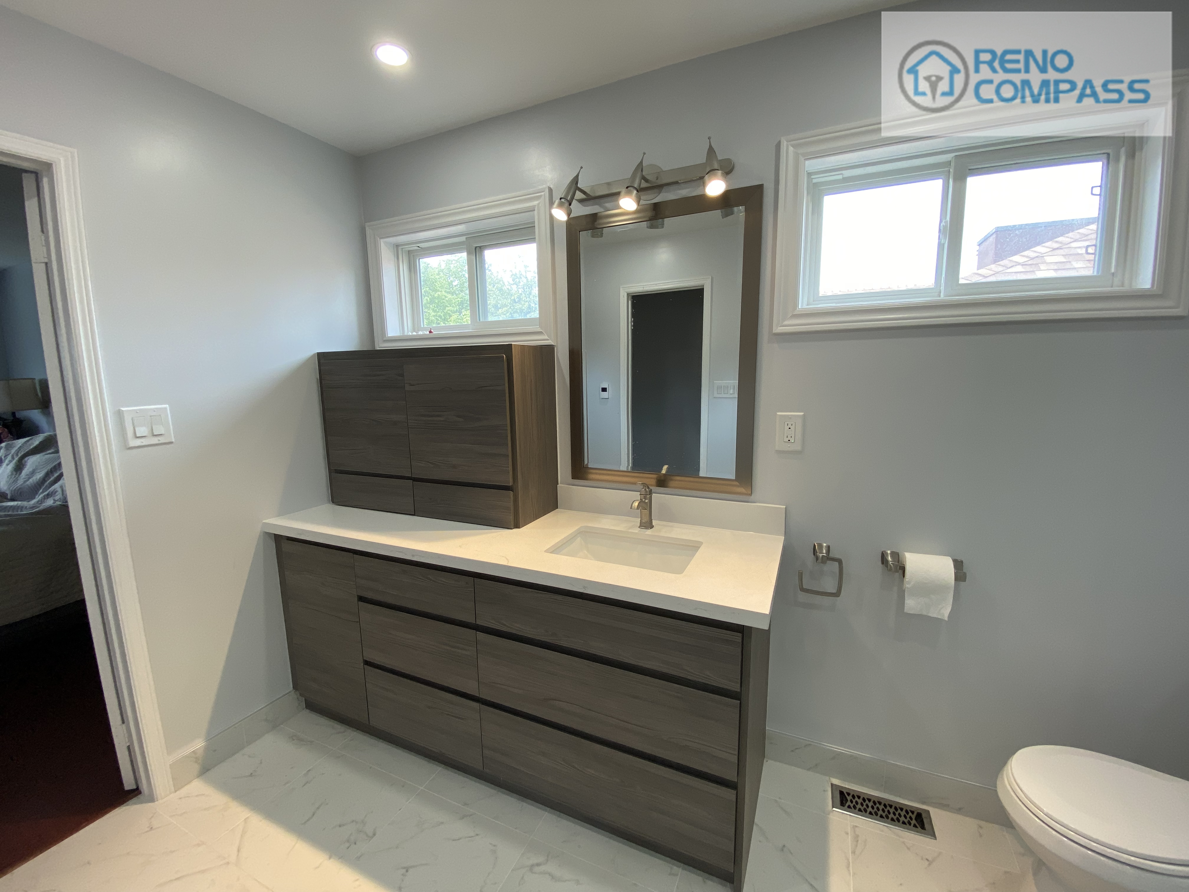 This bathroom renovation in Pickering showcases a modern wood-finish vanity with double drawers, a quartz countertop, integrated sink, and wall-mounted mirror with contemporary lighting. The space is enhanced by natural light from dual windows and a neutral color palette, creating a clean and practical bathroom environment. This project was completed by Reno Compass Inc. as part of a full interior renovation.
