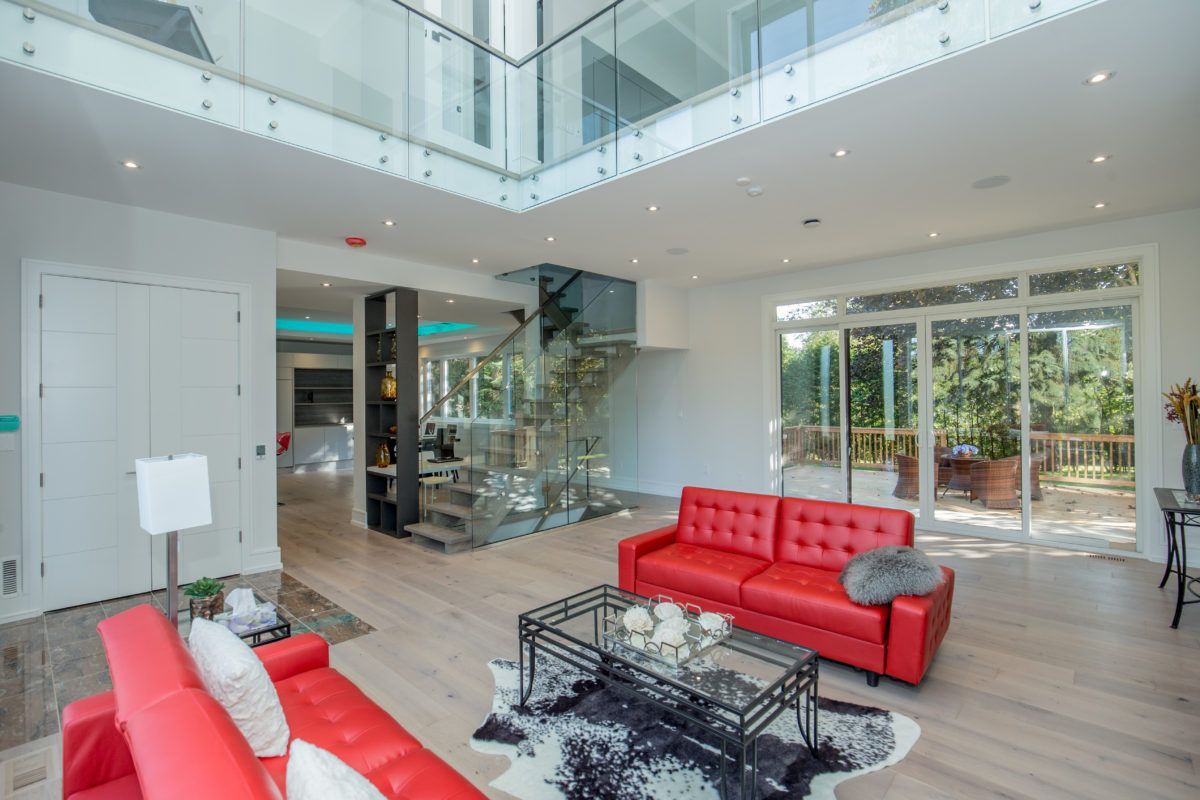Modern open-concept living room in a 5000 sqft Toronto home renovation featuring double-height ceilings, glass railing mezzanine, wide plank hardwood flooring, contemporary fireplace wall with built-in TV, and large sliding doors leading to a backyard deck. The space combines natural light, clean architectural lines, and bold red leather sofas to create a bright and sophisticated interior. Renovated by Reno Compass Inc., a professional Toronto renovation company specializing in large-scale custom home interior transformations.