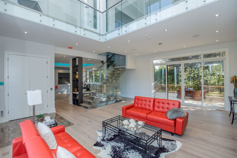 Modern open-concept living room in a 5000 sqft Toronto home renovation featuring double-height ceilings, glass railing mezzanine, wide plank hardwood flooring, contemporary fireplace wall with built-in TV, and large sliding doors leading to a backyard deck. The space combines natural light, clean architectural lines, and bold red leather sofas to create a bright and sophisticated interior. Renovated by Reno Compass Inc., a professional Toronto renovation company specializing in large-scale custom home interior transformations.
