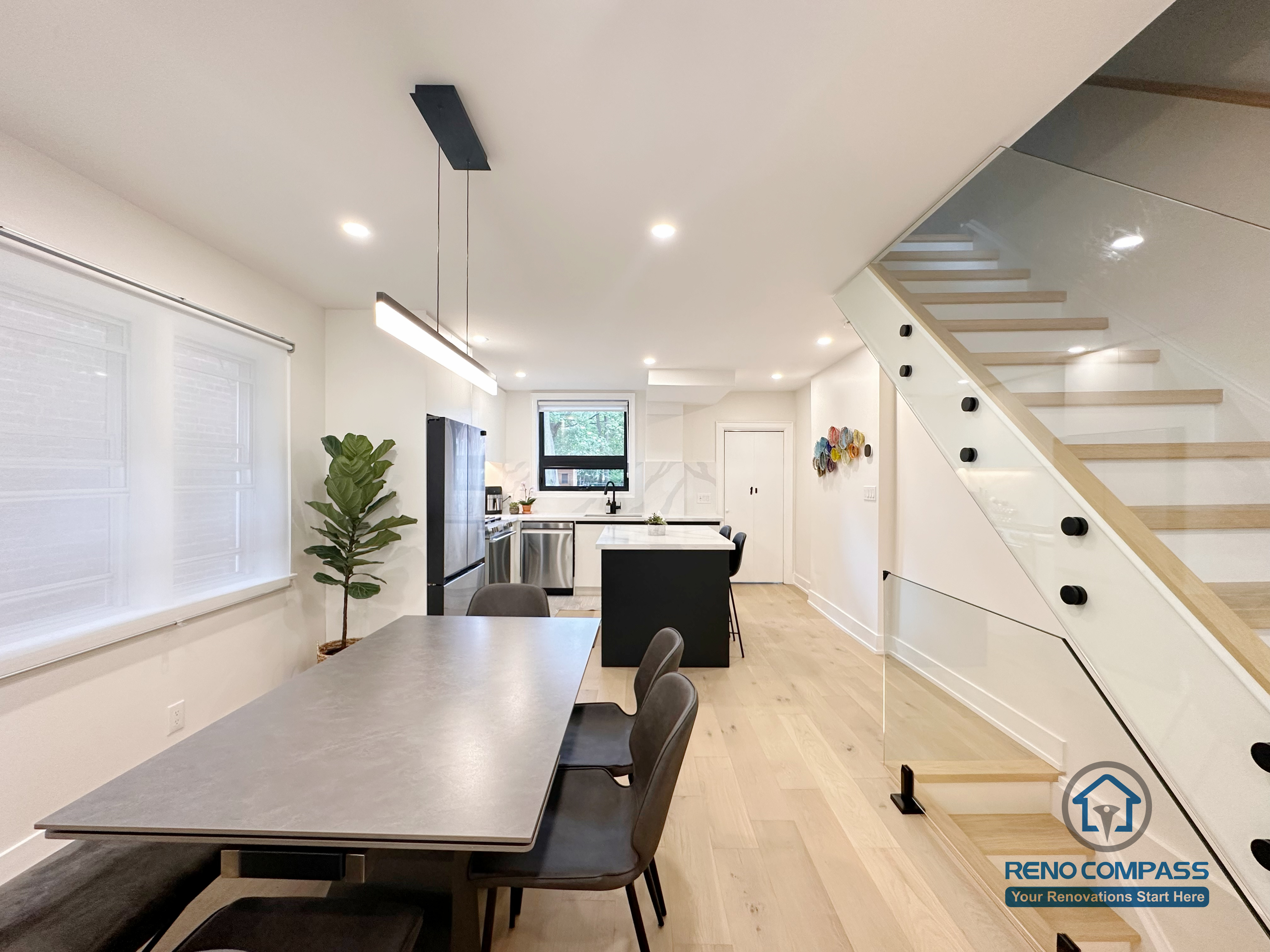 This Toronto interior renovation highlights a modern open-concept kitchen and dining area featuring a custom island, minimalist pendant lighting, wide-plank hardwood flooring, and a sleek glass staircase. The clean layout enhances natural light and flow, demonstrating refined craftsmanship by an experienced Toronto renovation company.