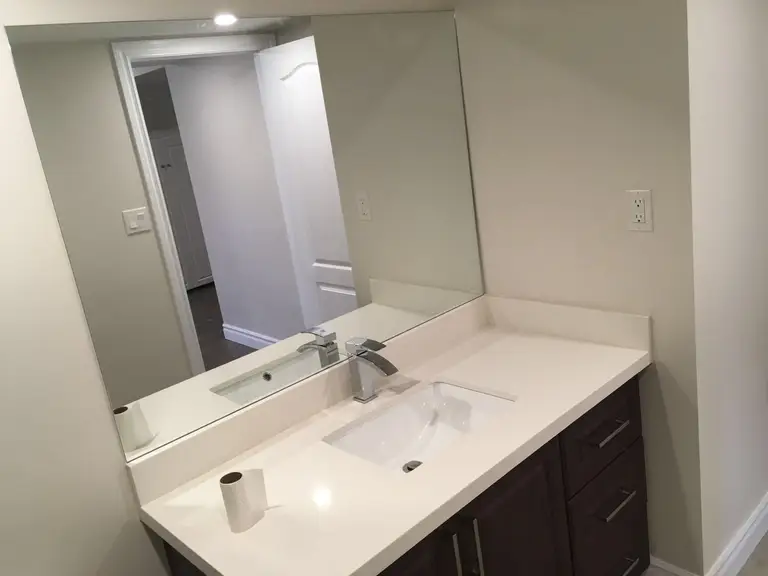 This Toronto interior renovation features a sleek bathroom vanity with a clean white quartz countertop, integrated sink, and a large frameless mirror. The design combines dark wood cabinetry with modern chrome fixtures, creating a simple yet elegant space that is both functional and easy to maintain. Neutral tones and efficient layout enhance brightness and usability.