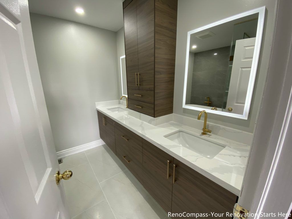 This Oakville master bathroom renovation showcases a modern double-sink vanity with custom wood cabinetry, quartz countertop, and elegant gold fixtures. The design features large-format floor tiles, LED backlit mirrors, and a sleek glass shower, creating a bright and luxurious spa-like atmosphere. Completed by a professional Toronto renovation company, this bathroom upgrade focuses on functionality, storage, and contemporary style, enhancing both comfort and property value for the homeowner.