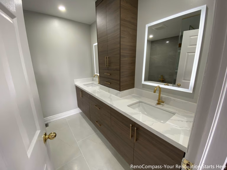 This Oakville master bathroom renovation showcases a modern double-sink vanity with custom wood cabinetry, quartz countertop, and elegant gold fixtures. The design features large-format floor tiles, LED backlit mirrors, and a sleek glass shower, creating a bright and luxurious spa-like atmosphere. Completed by a professional Toronto renovation company, this bathroom upgrade focuses on functionality, storage, and contemporary style, enhancing both comfort and property value for the homeowner.
