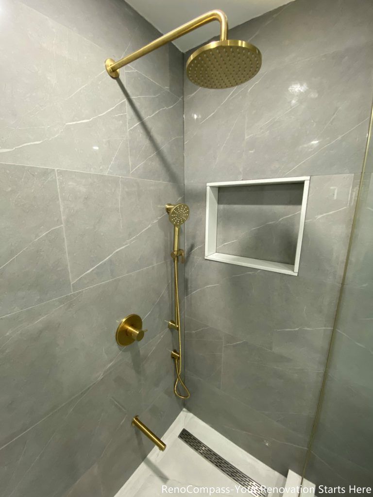 This Oakville master bathroom renovation features a sleek walk-in shower with premium gold rain shower system and modern handheld fixture for a luxury spa-like experience. Large-format marble-look wall tiles, a built-in recessed shower niche, and a linear floor drain create a clean, contemporary design with enhanced functionality. Completed by a professional Toronto renovation company, this bathroom upgrade combines elegant finishes, efficient drainage, and durable materials to deliver both comfort and long-term value for the homeowner.