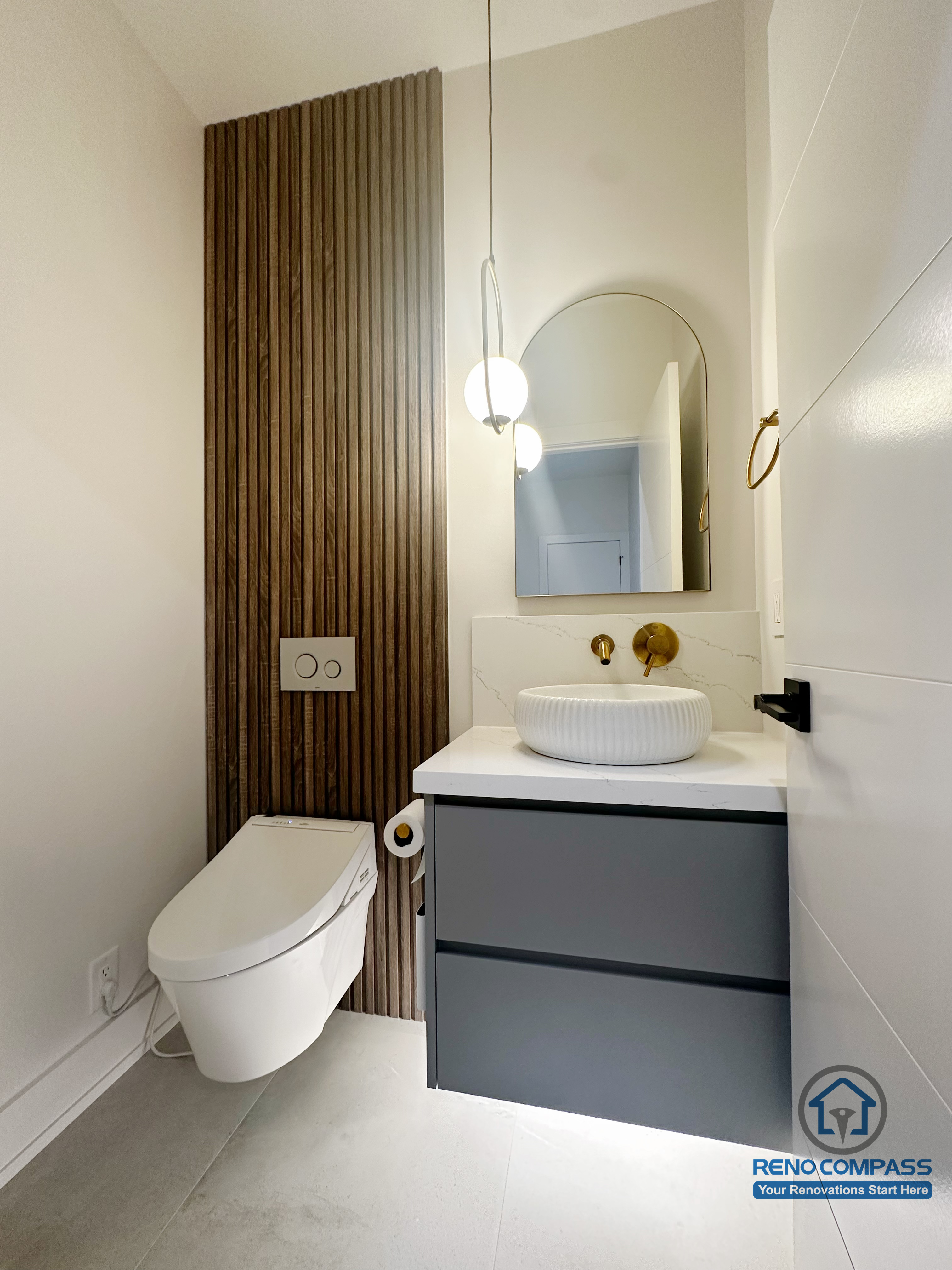 This Oakville powder room renovation highlights a compact yet elegant design with a floating vanity, vessel sink, and warm brass fixtures. The vertical wood slat feature wall adds visual depth and contrast, while the wall-mounted toilet and minimalist layout enhance openness, making the space both functional and visually refined.