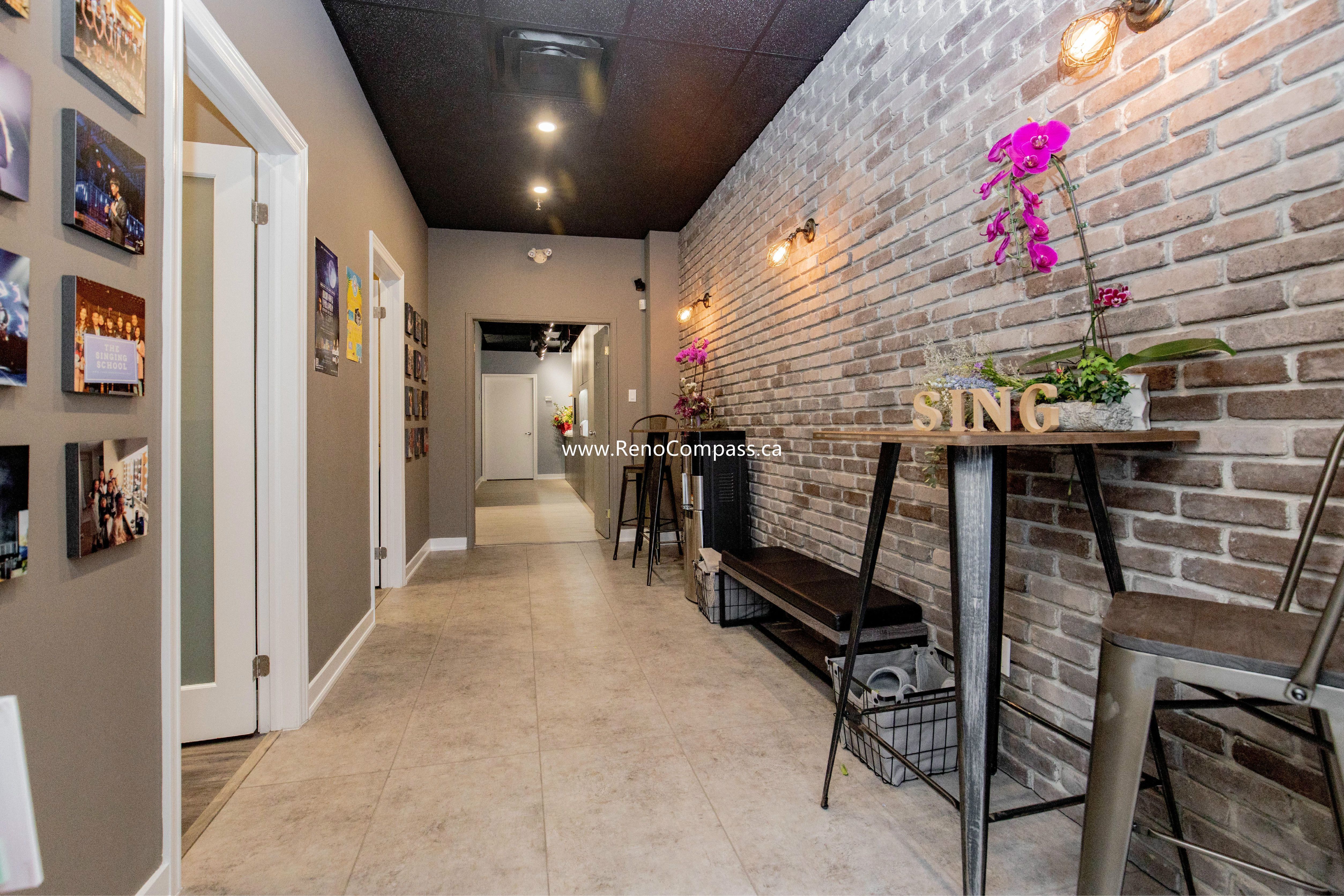 A professional commercial hallway transformation featuring a rustic brick veneer accent wall, industrial-style sconce lighting, and a modern black drop ceiling. This Richmond Hill project for The Singing School showcases functional, stylish circulation space with high-traffic tile flooring.