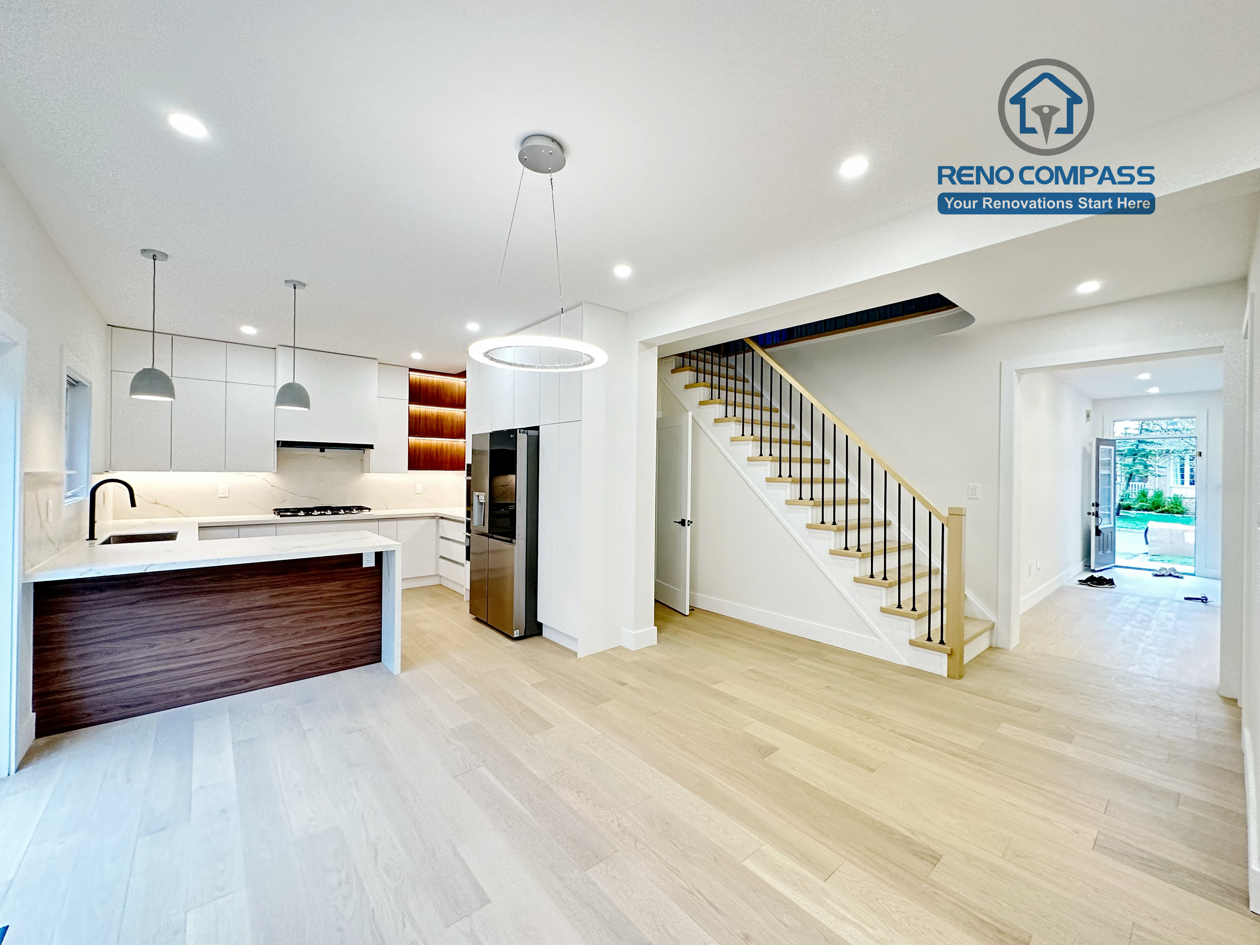 This image showcases a modern open-concept kitchen renovation completed in Richmond Hill. The project features custom white cabinetry, a large waterfall kitchen island with wood panel detailing, built-in stainless steel appliances, and seamless integration with the dining and staircase area. Bright recessed lighting, minimalist pendant lights, and light hardwood flooring enhance the clean and spacious feel of the home. Completed by a professional Toronto Renovation Company, this kitchen renovation combines functionality, contemporary design, and high-quality craftsmanship tailored to modern family living.