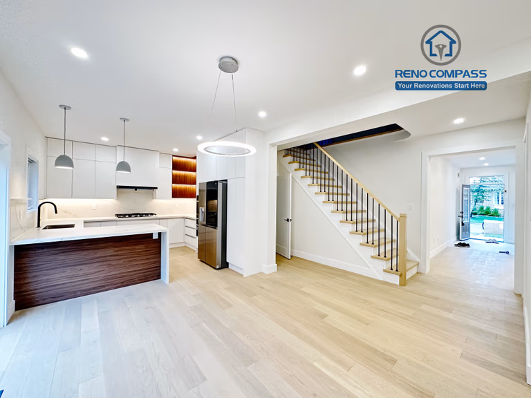 This image showcases a modern open-concept kitchen renovation completed in Richmond Hill. The project features custom white cabinetry, a large waterfall kitchen island with wood panel detailing, built-in stainless steel appliances, and seamless integration with the dining and staircase area. Bright recessed lighting, minimalist pendant lights, and light hardwood flooring enhance the clean and spacious feel of the home. Completed by a professional Toronto Renovation Company, this kitchen renovation combines functionality, contemporary design, and high-quality craftsmanship tailored to modern family living.