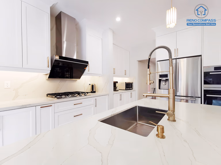 This Markham interior renovation highlights a custom kitchen island designed as a central functional workspace within the home. The oversized quartz countertop provides generous prep and serving space, while the integrated undermount sink and professional pull-down faucet enhance daily usability. White shaker cabinetry paired with warm metal hardware creates a balanced contrast between clean structure and subtle elegance. Built-in appliances and a streamlined range hood maintain visual clarity and efficient workflow across the kitchen. Completed by Reno Compass, this kitchen renovation reflects careful attention to proportion, material selection, and long-term practicality, tailored to modern Markham homeowners seeking refined yet highly functional interior design.