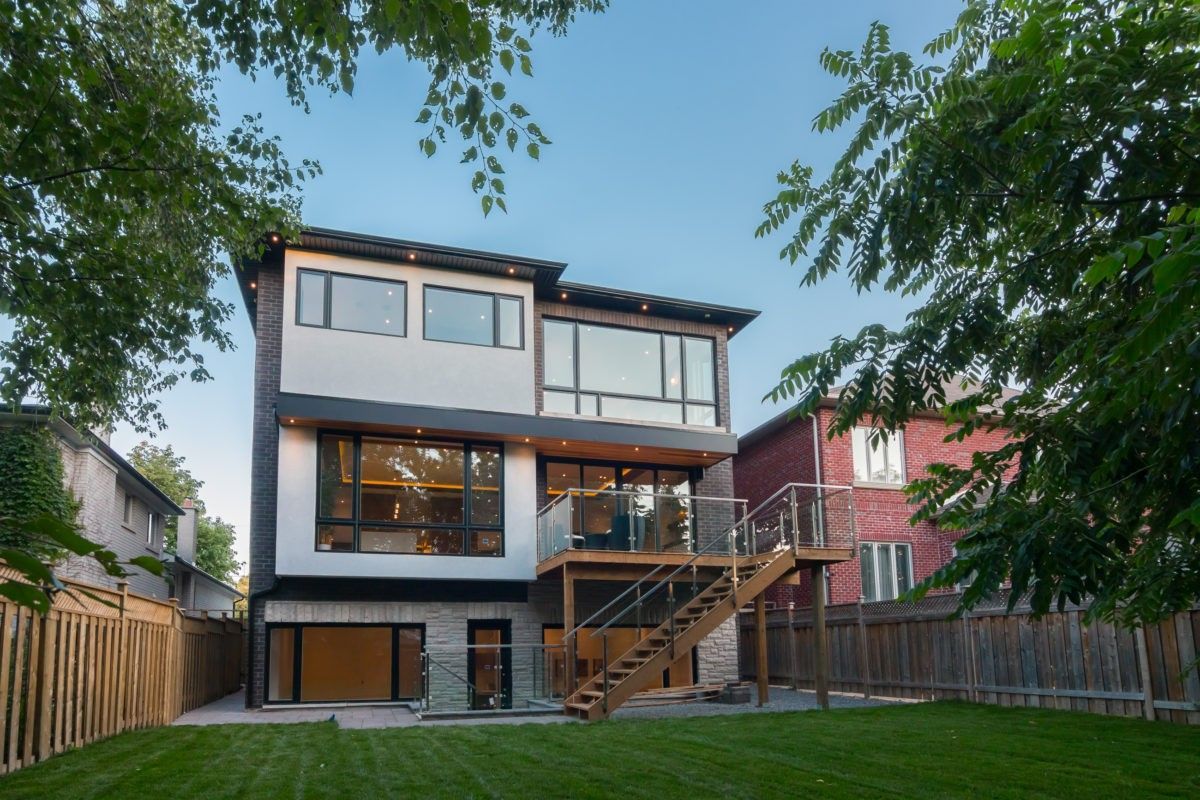 This Toronto modern home renovation showcases a stunning rear exterior featuring a raised wood deck with glass railings, large windows, and a seamless connection between indoor and outdoor living spaces. The clean architectural lines, modern materials, and landscaped backyard create a functional and visually striking contemporary home design.