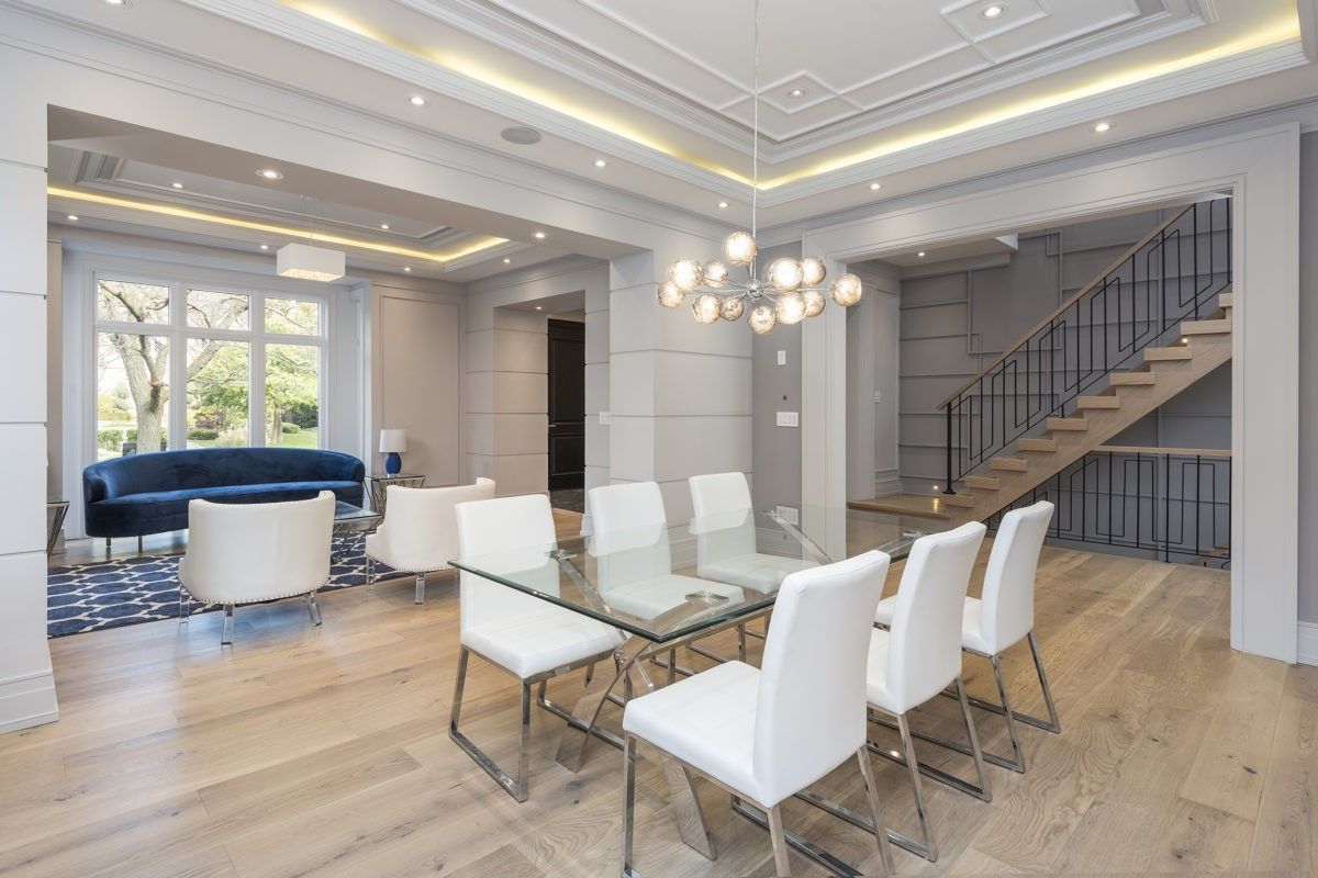 This Newmarket interior renovation highlights a modern luxury dining room completed by a professional Toronto renovation company. The space features wide plank hardwood flooring, custom wall panel detailing, and a coffered ceiling with warm recessed LED lighting that enhances depth and elegance. A contemporary glass dining table with upholstered chairs and a designer chandelier creates a refined focal point, while large windows bring in natural light. The renovation delivers a sophisticated open-concept dining area ideal for modern family living and entertaining in Newmarket and across the GTA.