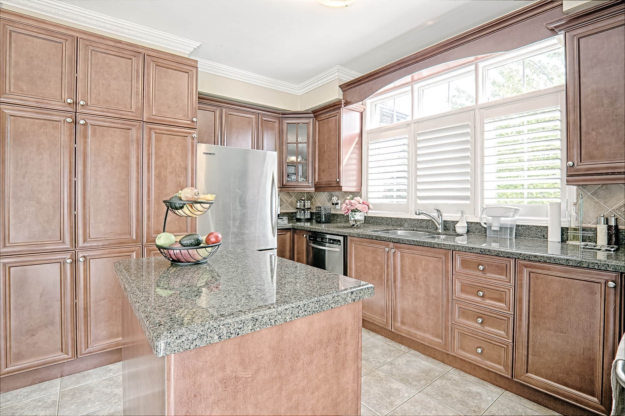 This warm traditional kitchen features rich wood cabinetry, granite countertops, and a functional center island that enhances both storage and workspace. Large windows with plantation shutters bring in natural light, creating a bright and inviting cooking environment. Stainless steel appliances and classic finishes blend timeless style with everyday practicality. A perfect example of elegant and functional kitchen renovation design for homeowners in Toronto and the GTA.