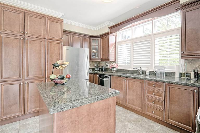 This warm traditional kitchen features rich wood cabinetry, granite countertops, and a functional center island that enhances both storage and workspace. Large windows with plantation shutters bring in natural light, creating a bright and inviting cooking environment. Stainless steel appliances and classic finishes blend timeless style with everyday practicality. A perfect example of elegant and functional kitchen renovation design for homeowners in Toronto and the GTA.
